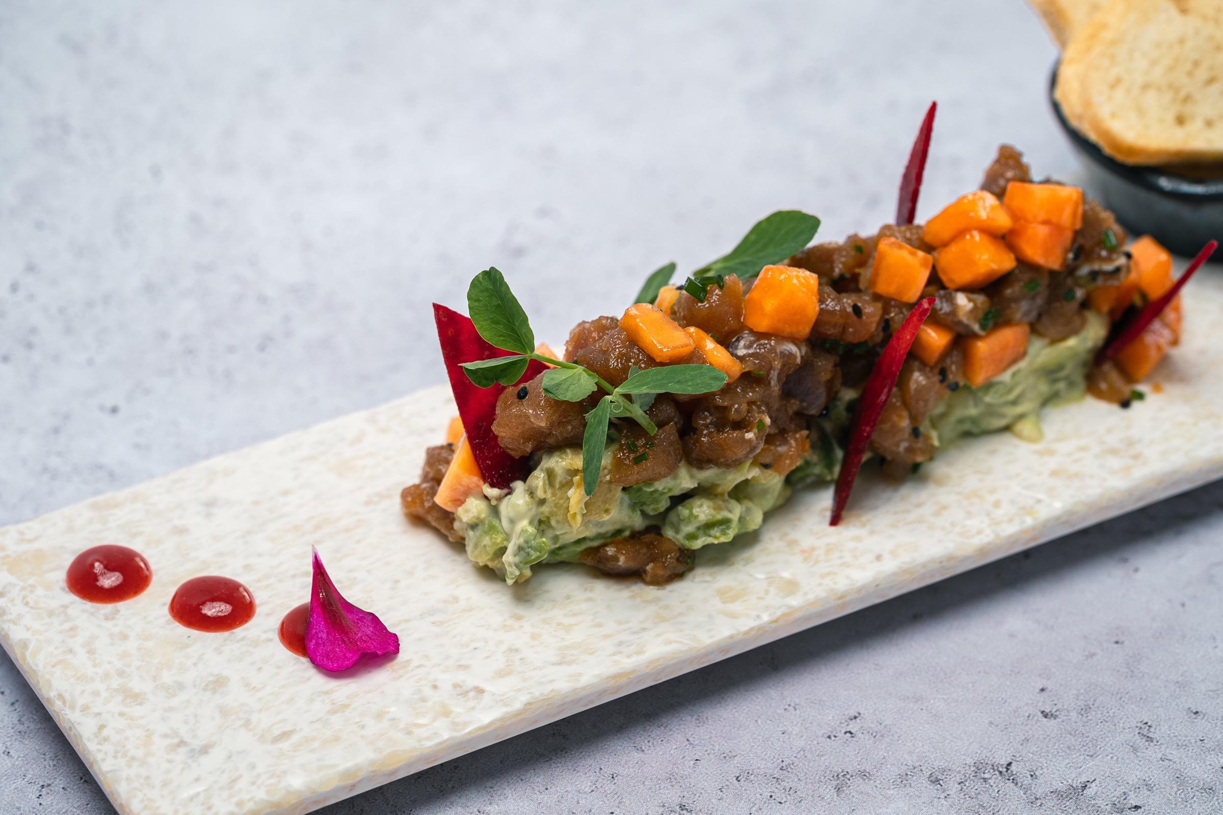 A tuna tartare plated in a long strip on a marble board, topped with diced mango, herbs and a beetroot crisp, with sauce dots and a flower petal beside it.