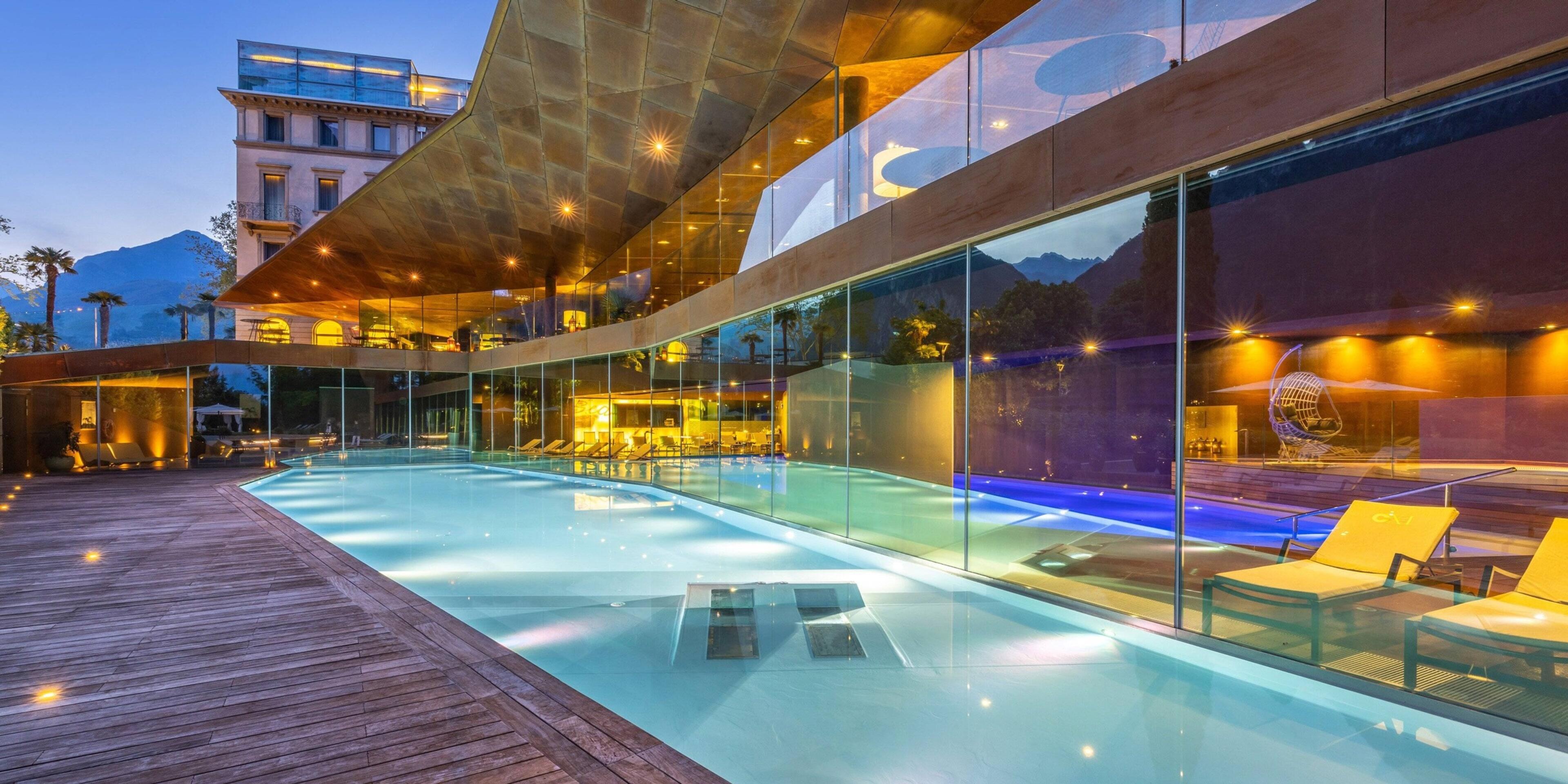 An outdoor infinity pool lit blue at dusk, running beside a glass-walled wing of the hotel with mountains behind.