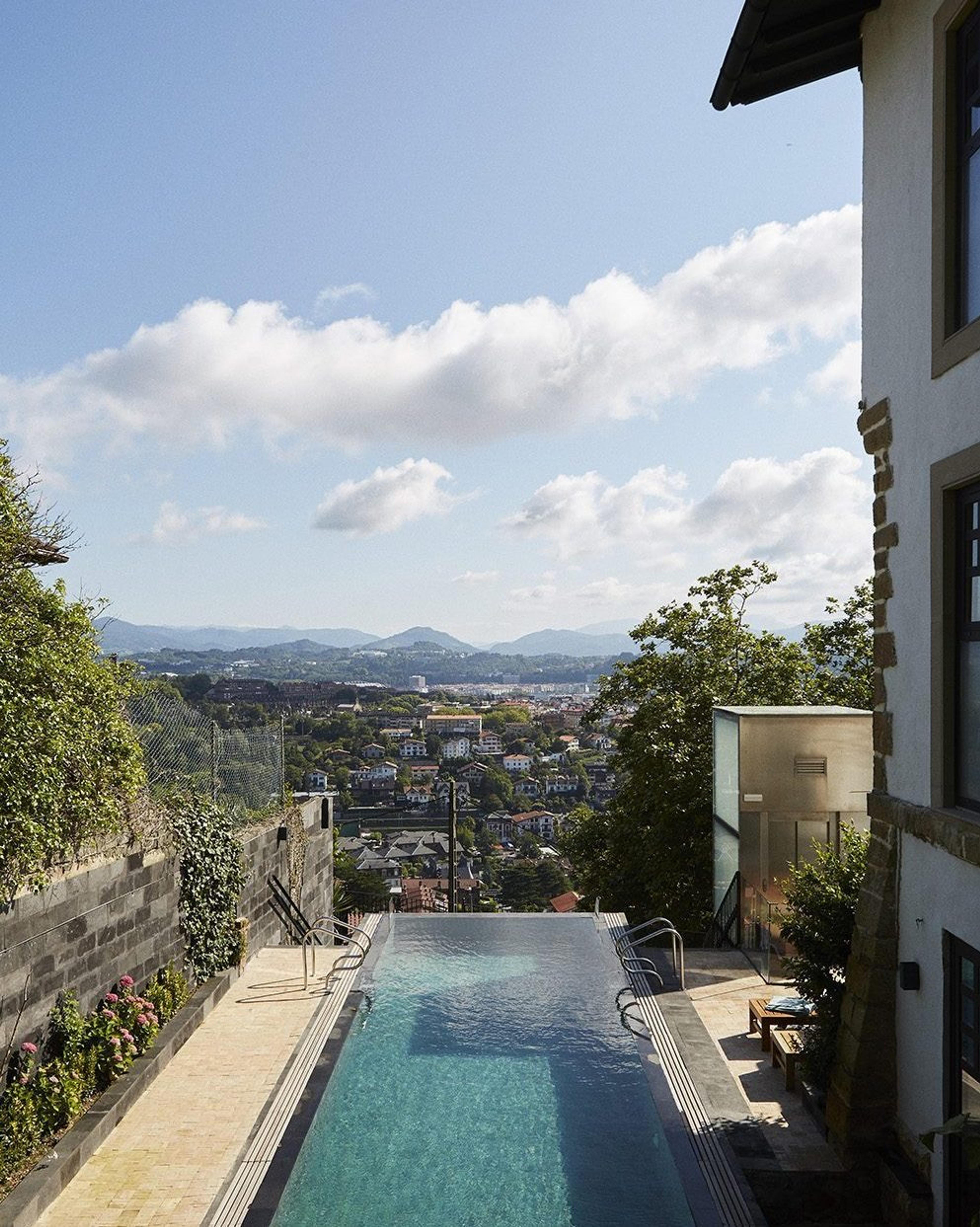 an infinity pool running along a terrace with a glazed structure and sun loungers beside it, overlooking the town, bay and hills