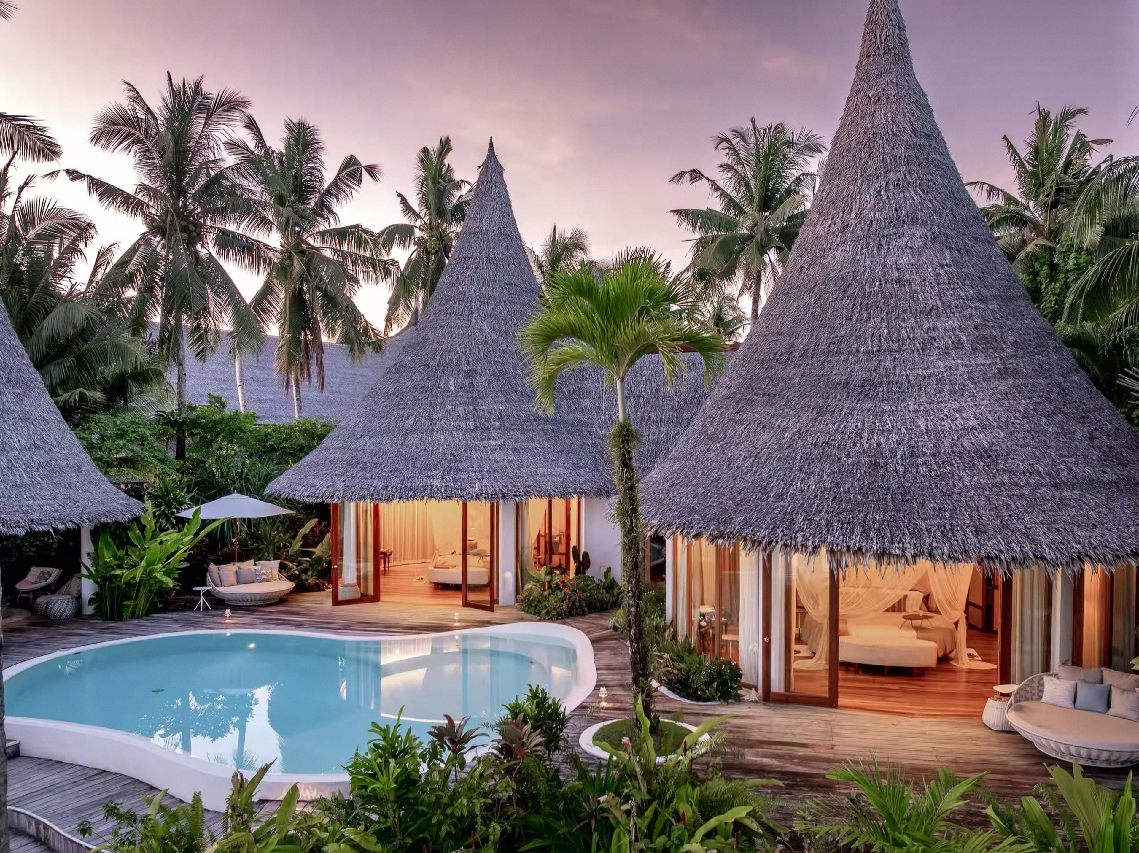 Two thatched roof villas at dusk with a curved pool in front and lit bedrooms visible through open doors.