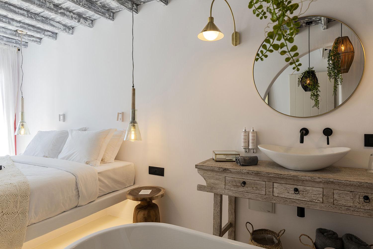 A bedroom and ensuite bathroom under a beamed ceiling, with a round mirror over a freestanding vessel sink and the bed visible alongside.