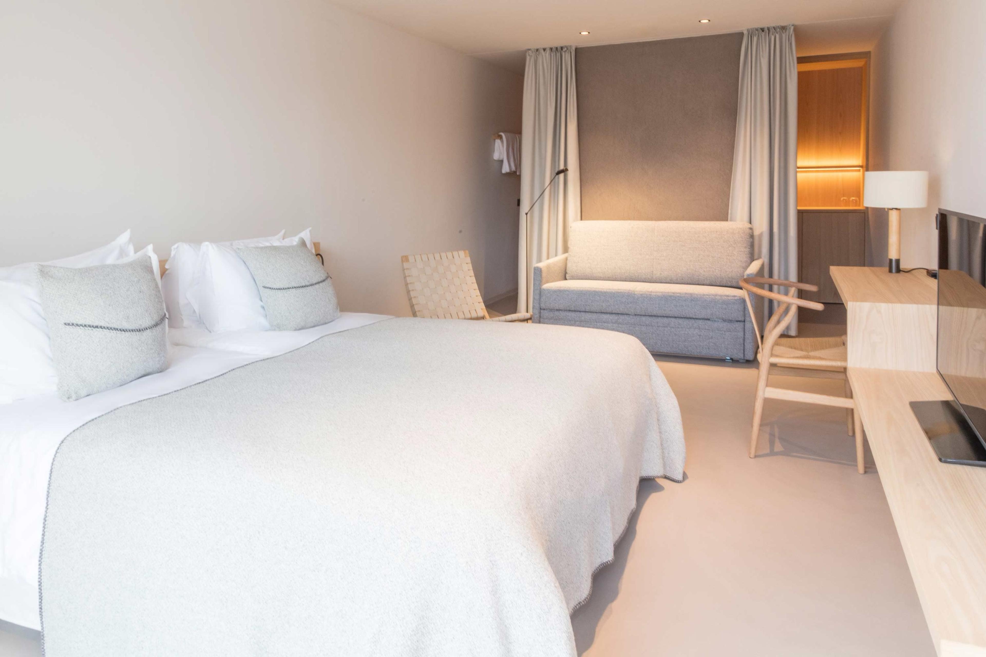 A hotel bedroom with a built-in grey upholstered bench between curtains, and a wooden desk with a lamp and an open laptop.