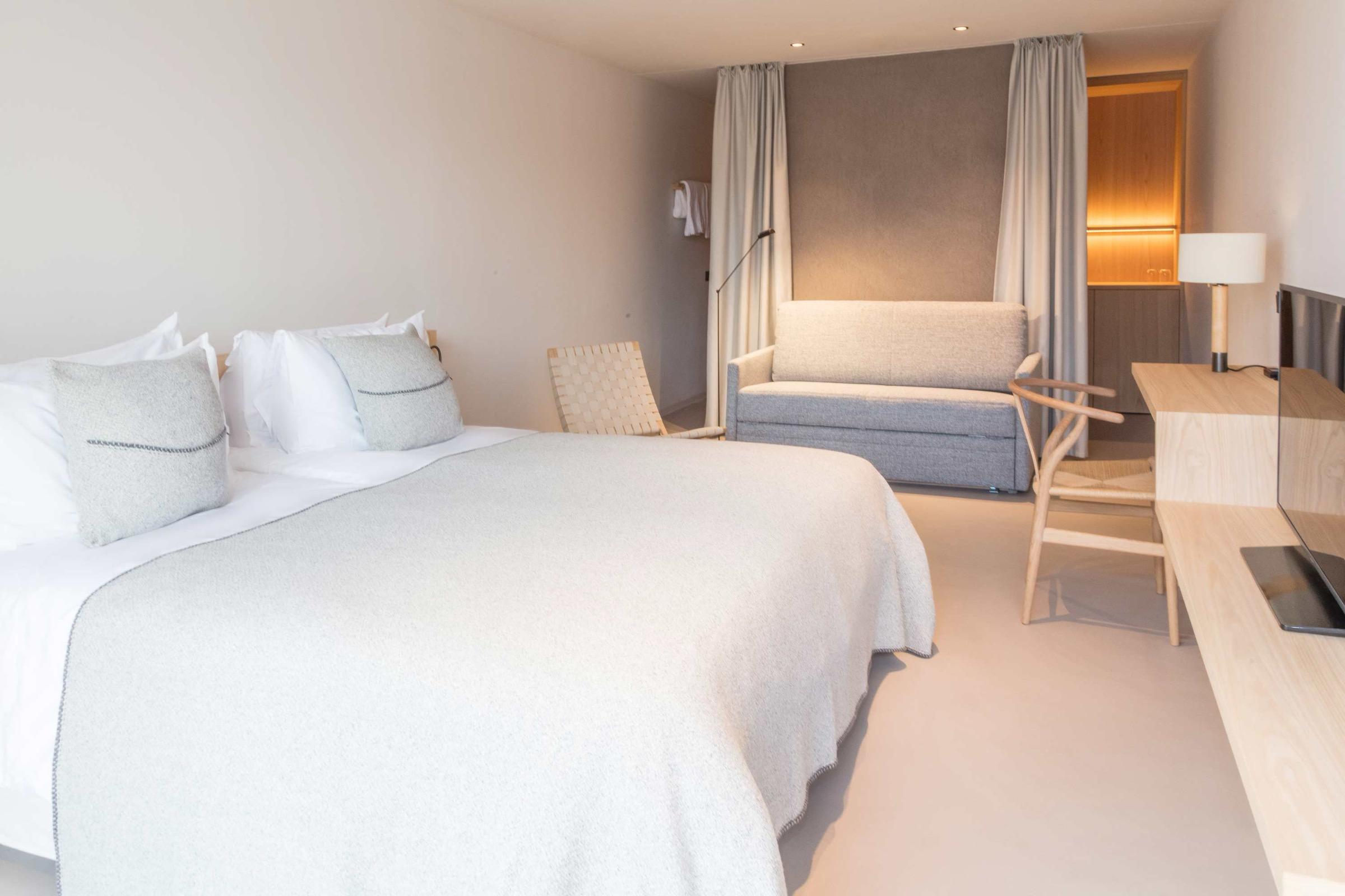 A hotel bedroom with a built-in grey upholstered bench between curtains, and a wooden desk with a lamp and an open laptop.
