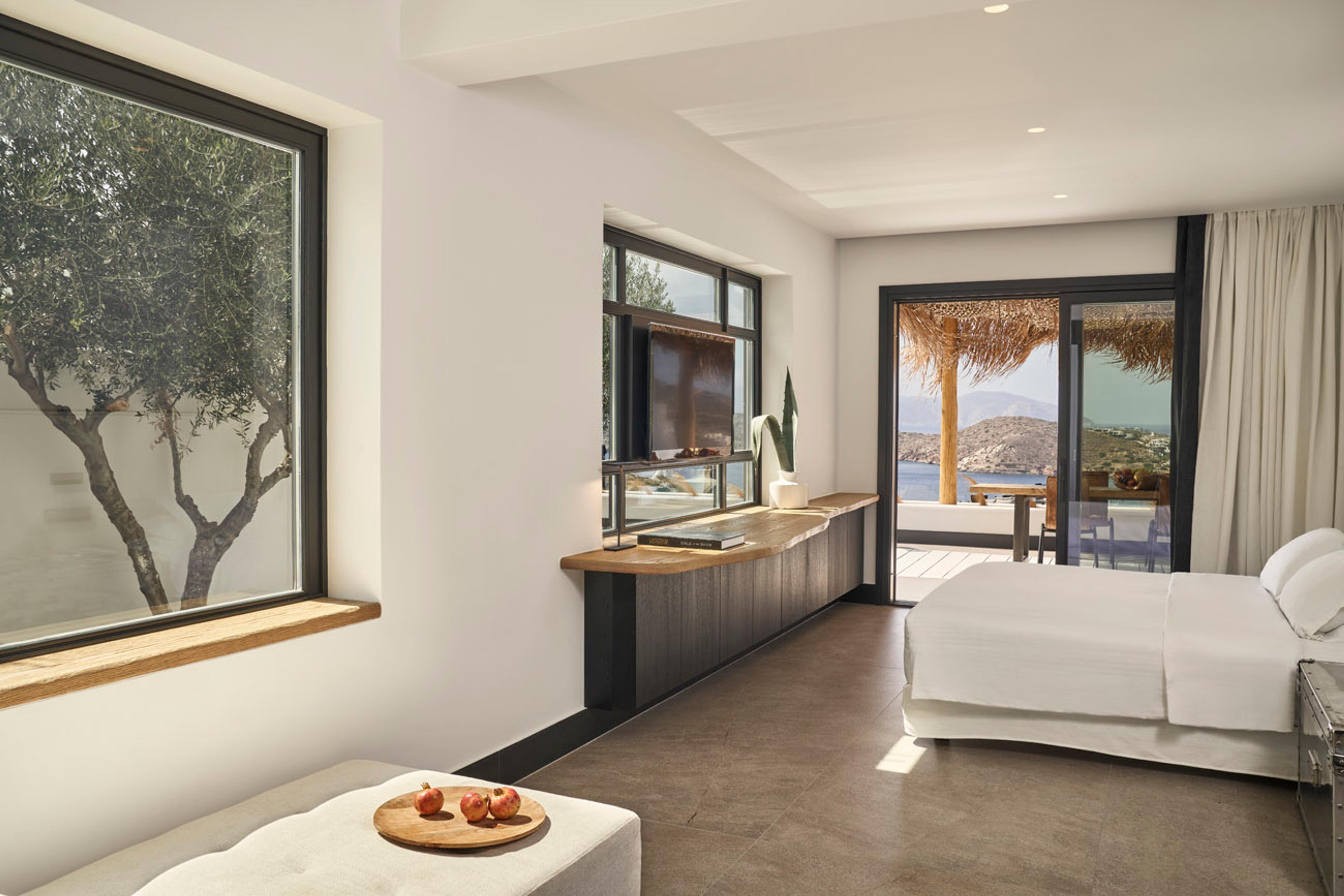 A bedroom with a large window framing an olive tree, a wooden ledge holding a television, and glass doors to a thatched terrace with the sea beyond.