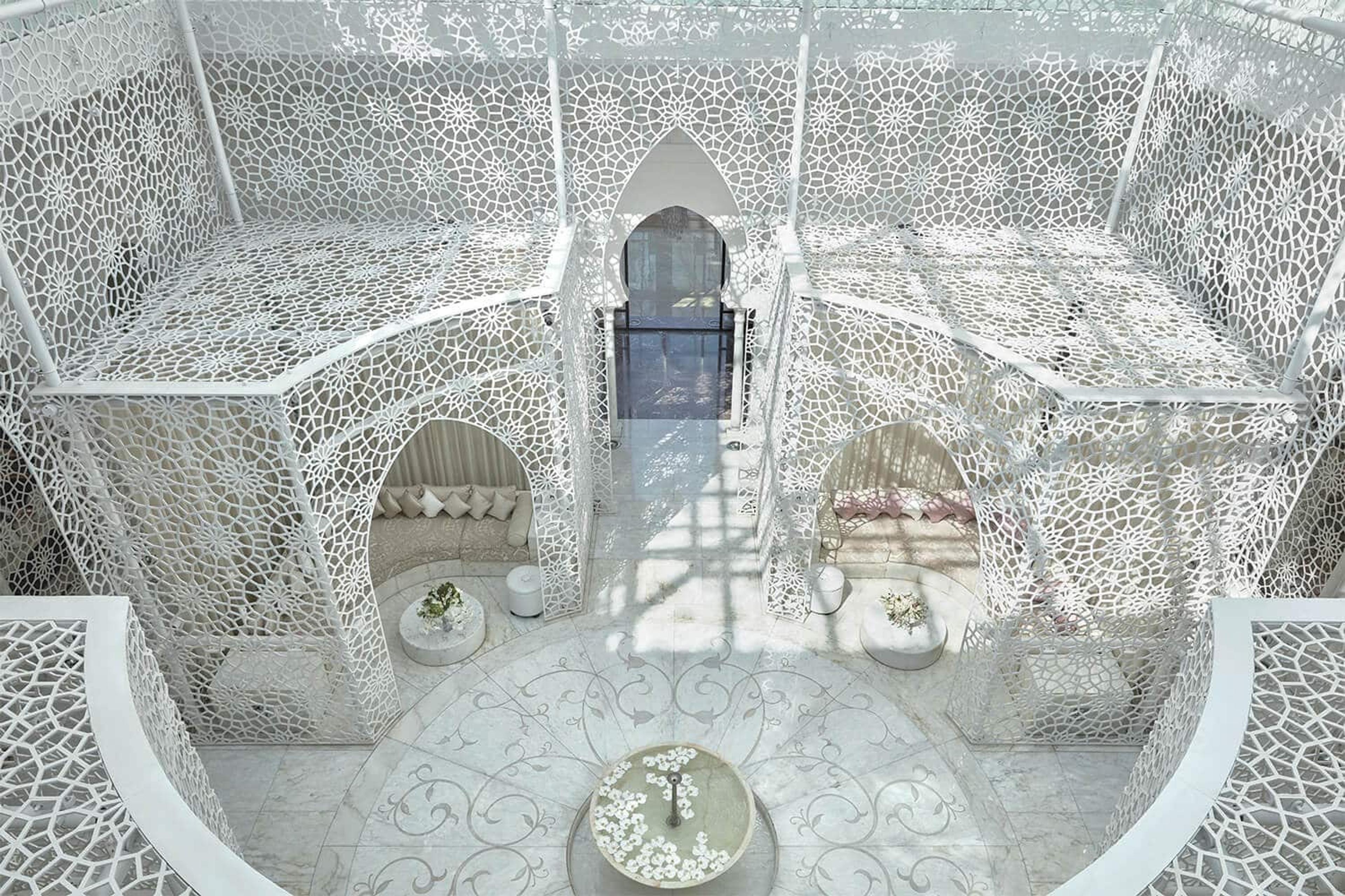 An overhead view of a white latticed courtyard with alcove seating areas and a central fountain.