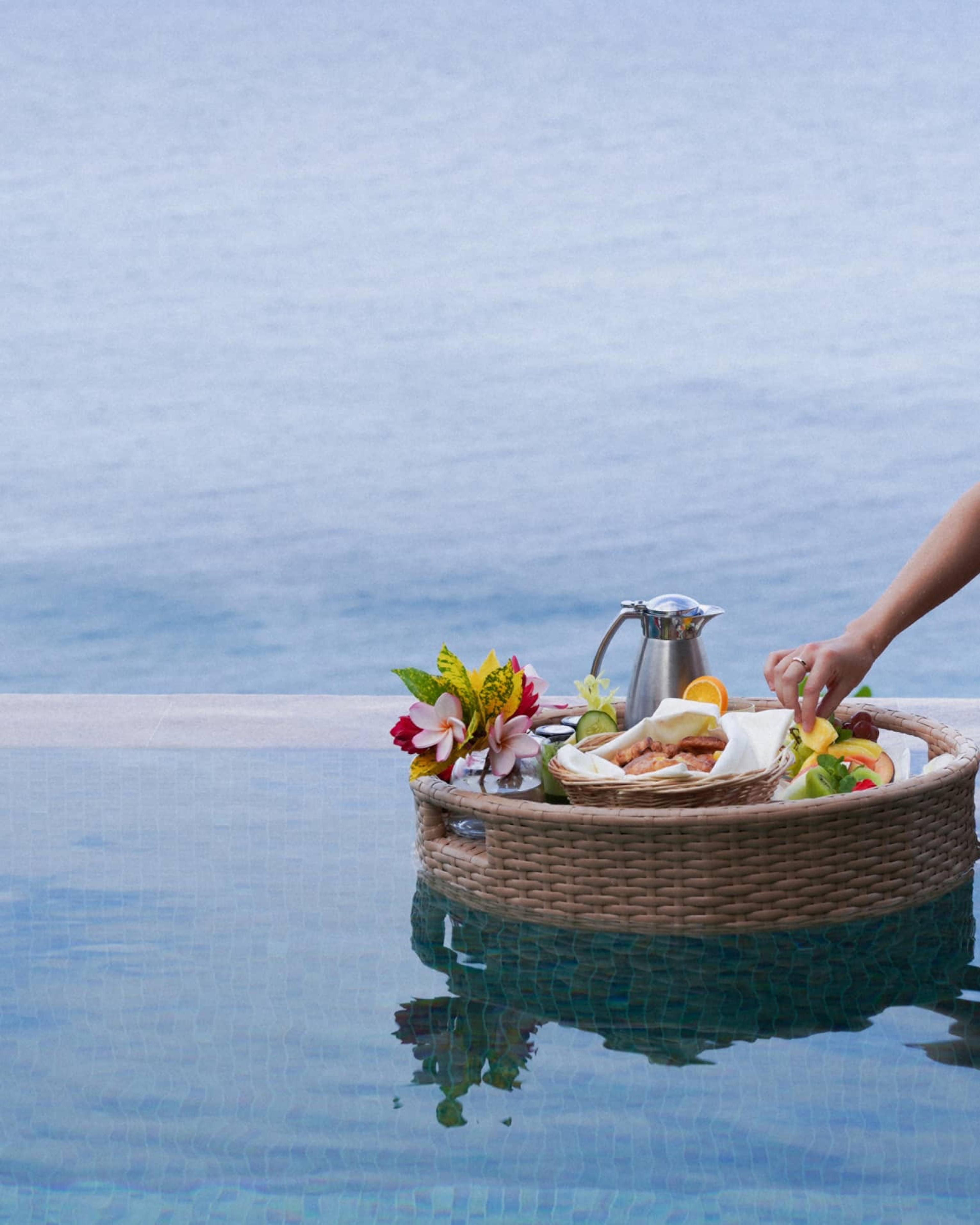 A hand reaching into a floating wicker tray of fruit, pastries and flowers on a pool, with the sea beyond.