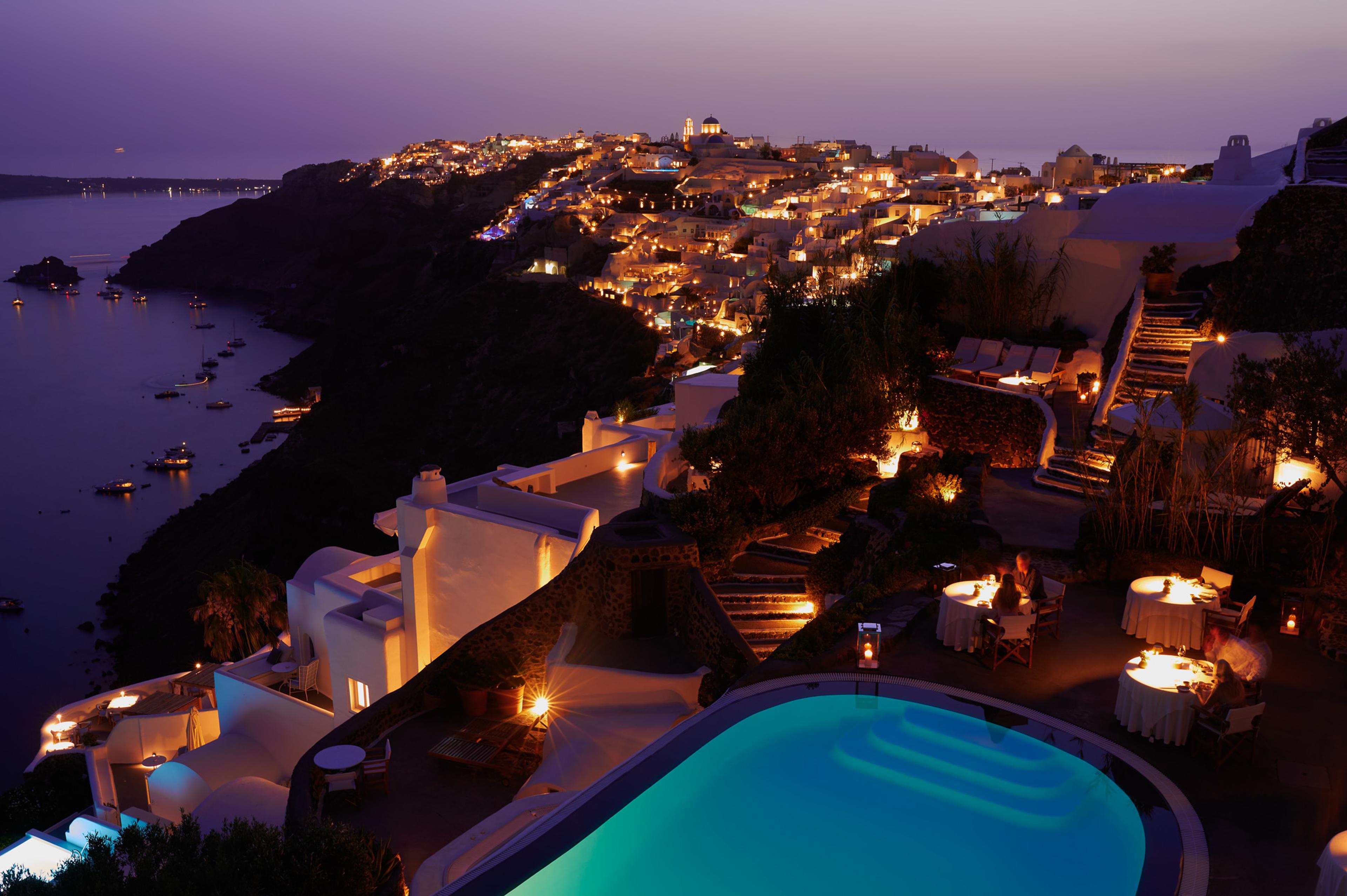 An aerial view at dusk of a hotel built into a cliffside above the sea, its lit pool and terraces in the foreground, diners at candlelit tables, and a clifftop village glowing across the water.