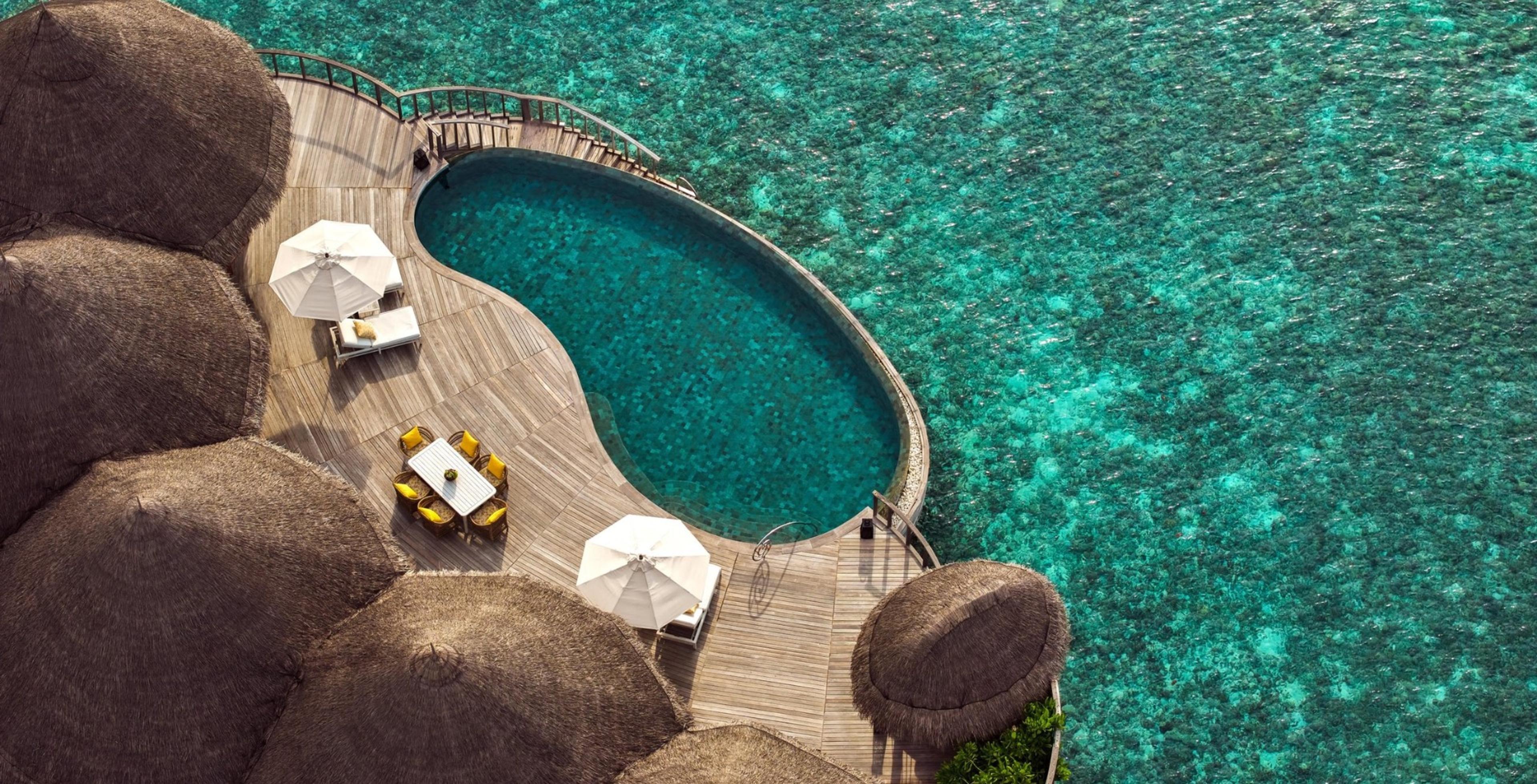 A top-down aerial view of an overwater pool deck with sun umbrellas, loungers and a dining table, set against a coral reef.