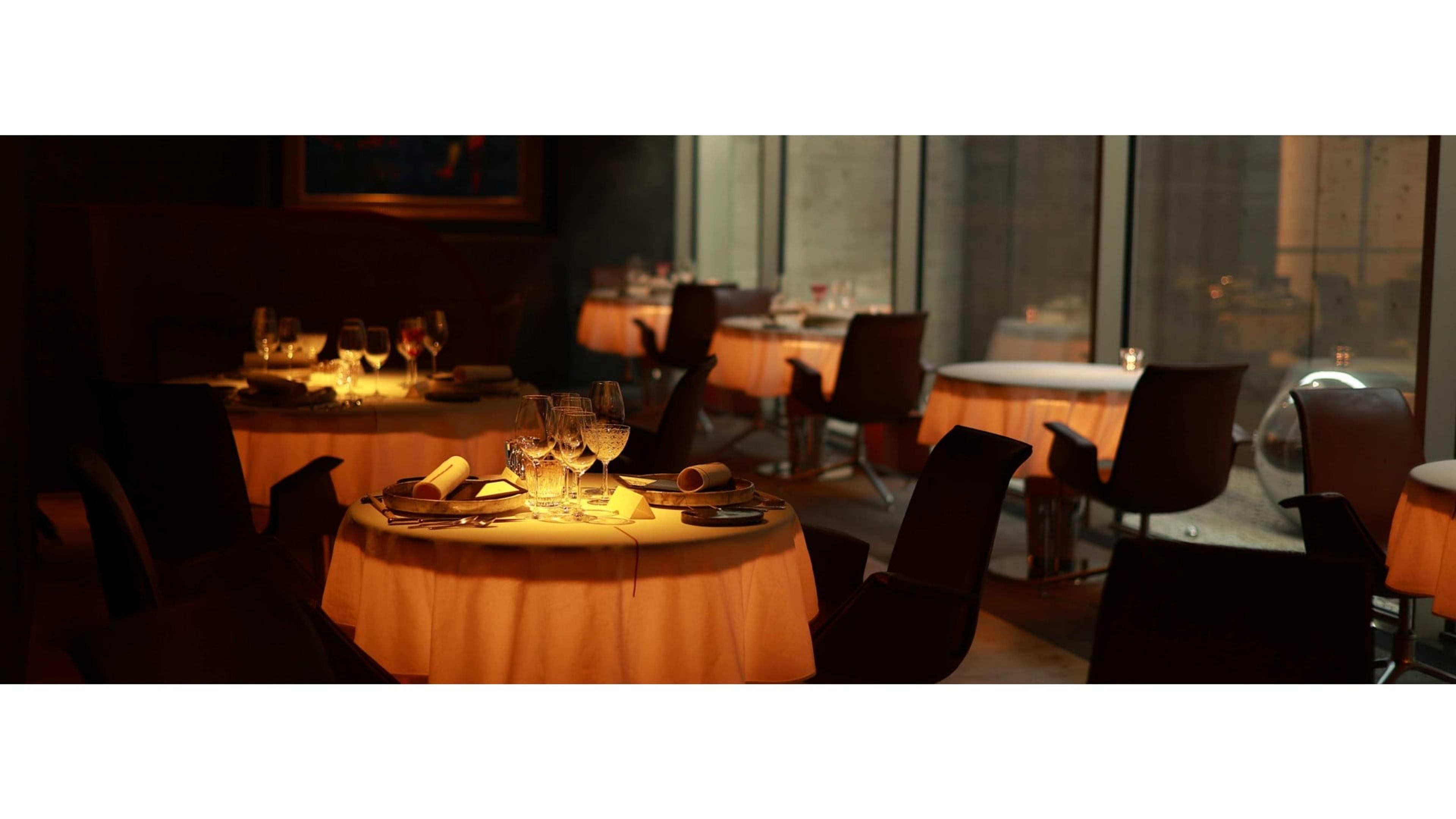 A dimly lit restaurant with round tables set for dinner and empty chairs, floor-to-ceiling windows along one side