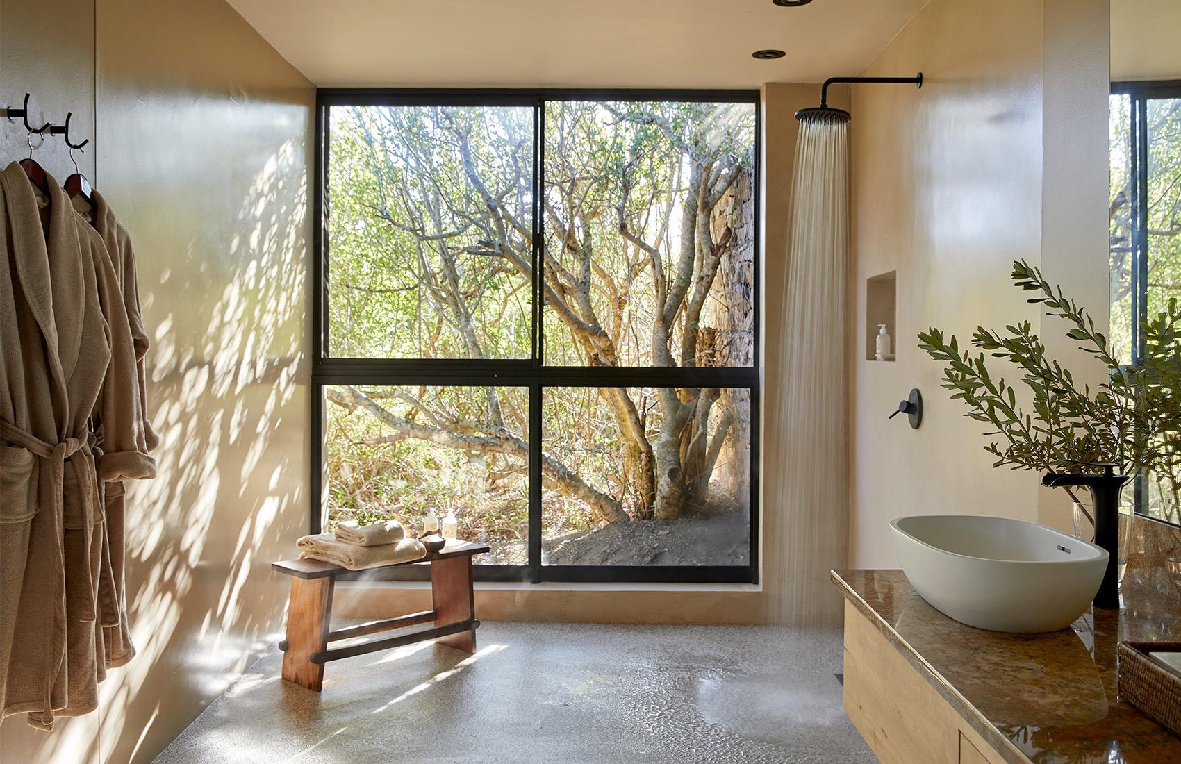 A bathroom with a freestanding basin, a walk-in shower, and a large window looking onto trees, with robes hanging on hooks.