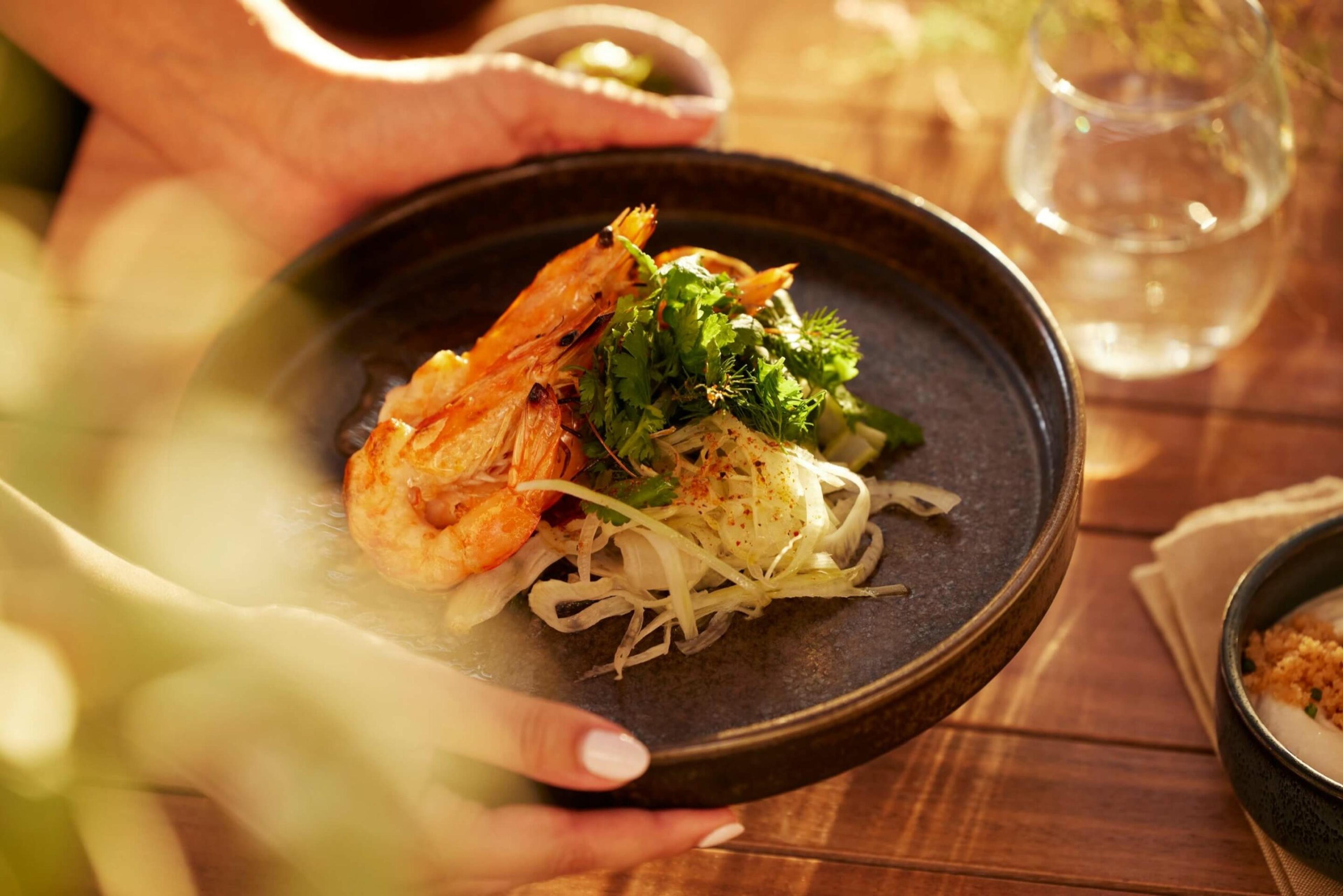 Hands hold out a plate of grilled prawns on a herb and fennel salad, set on a wooden table with a glass of water and blurred leaves in the foreground.