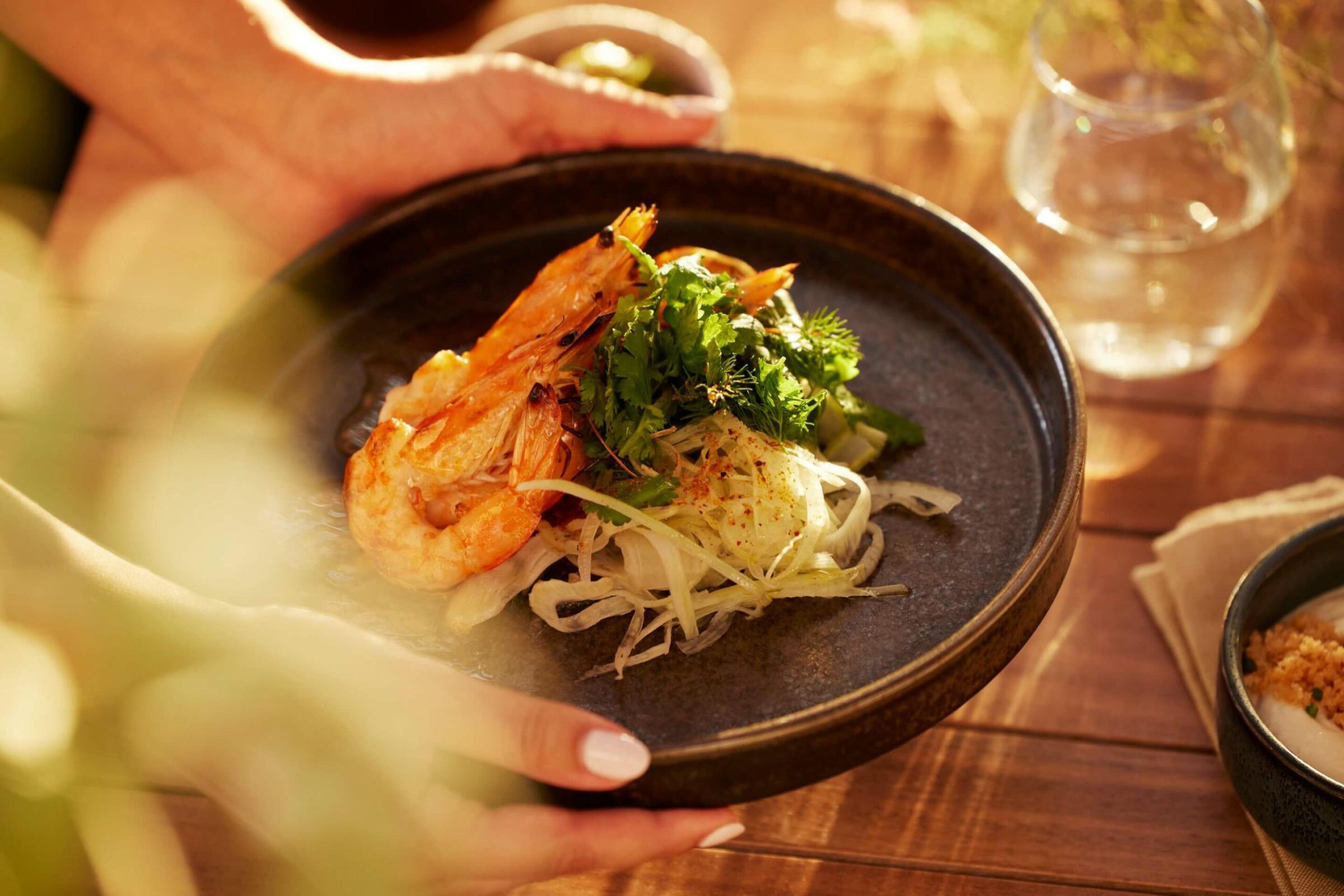 Hands hold out a plate of grilled prawns on a herb and fennel salad, set on a wooden table with a glass of water and blurred leaves in the foreground.