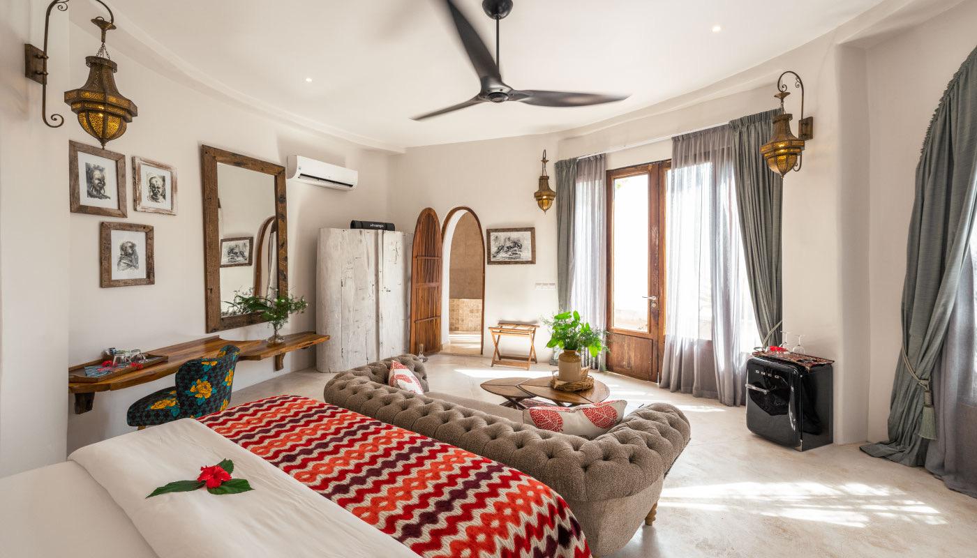 A bedroom with a patterned throw on the bed, a mirror and framed prints on the wall, and a ceiling fan overhead.