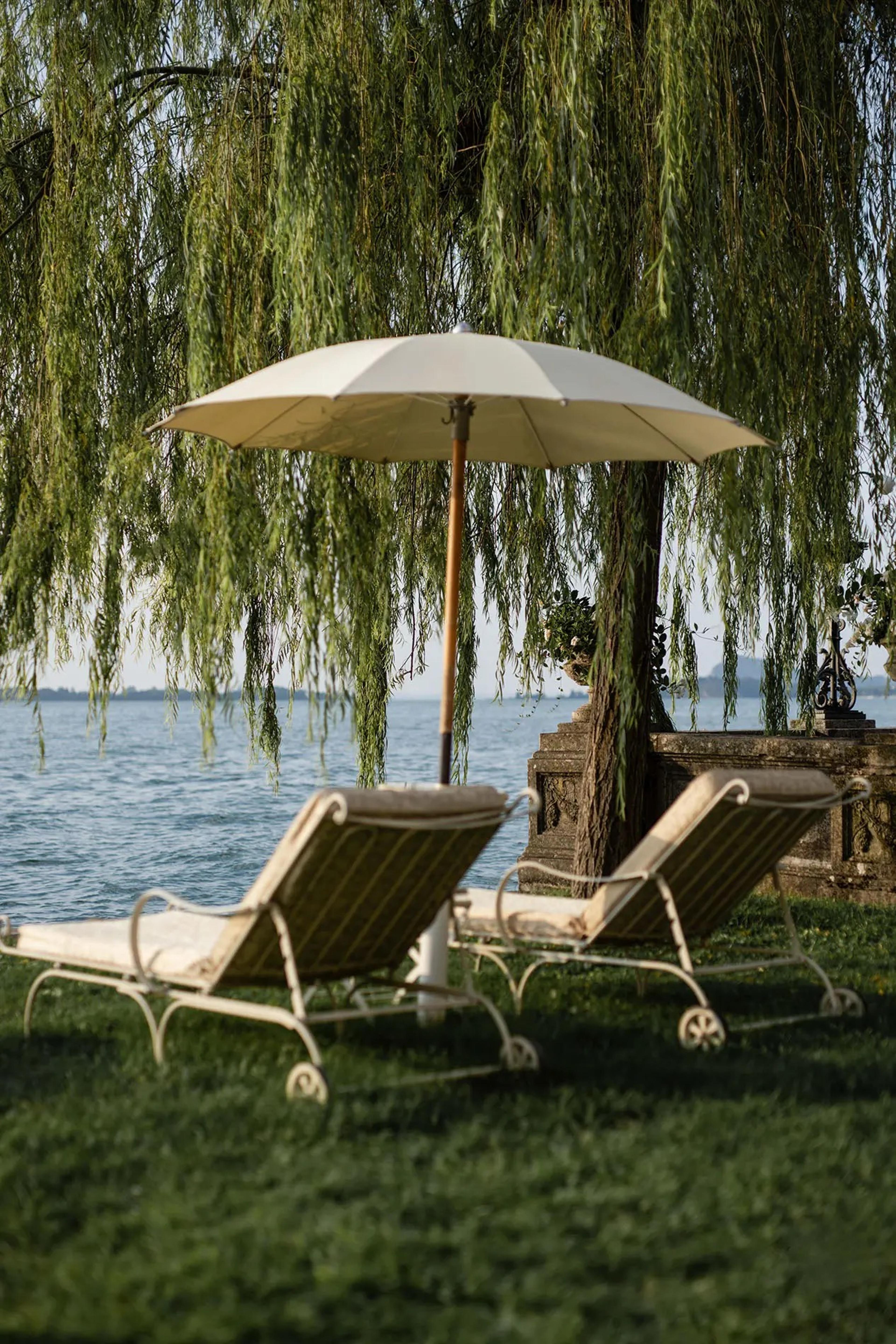 Two cushioned sun loungers and a folded white umbrella sit on grass under a weeping willow, facing an open lake.