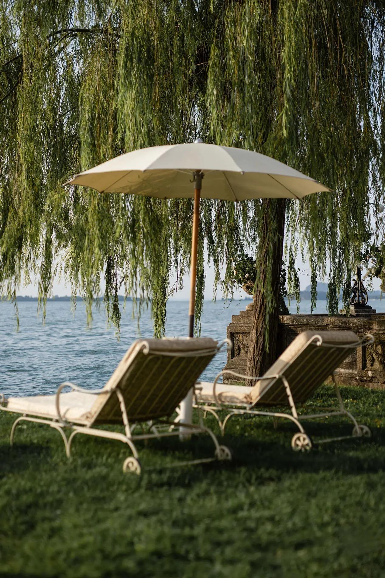 Two cushioned sun loungers and a folded white umbrella sit on grass under a weeping willow, facing an open lake.