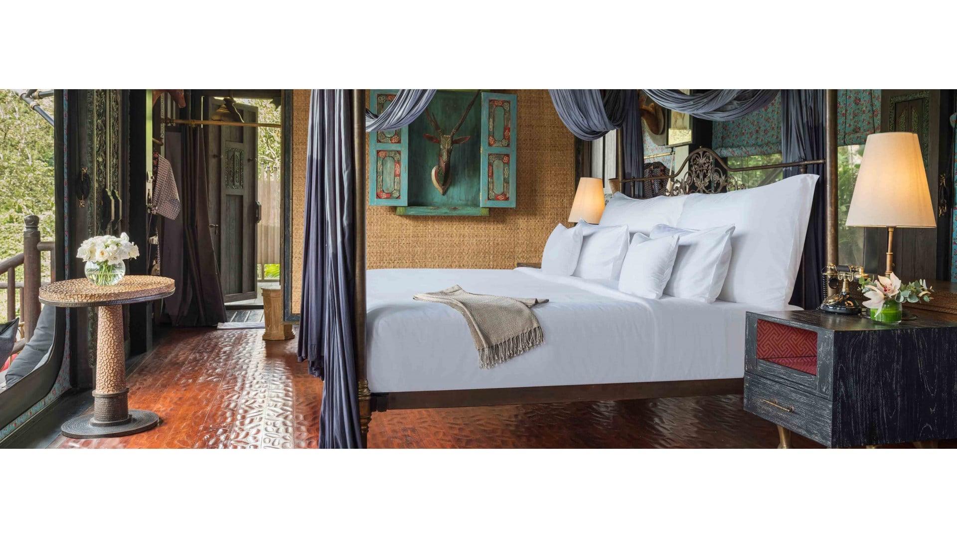A four-poster bed with white linen and dark drapes in a room with dark wood floors, a round table with flowers to the left and an open doorway showing trees beyond.