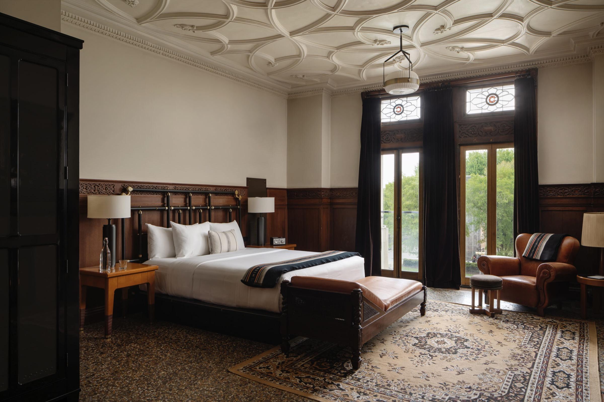 A king bed with a carved dark wood headboard beneath an ornate plaster ceiling, with stained glass topped French doors open to a balcony and trees, a wardrobe, and a leather armchair.