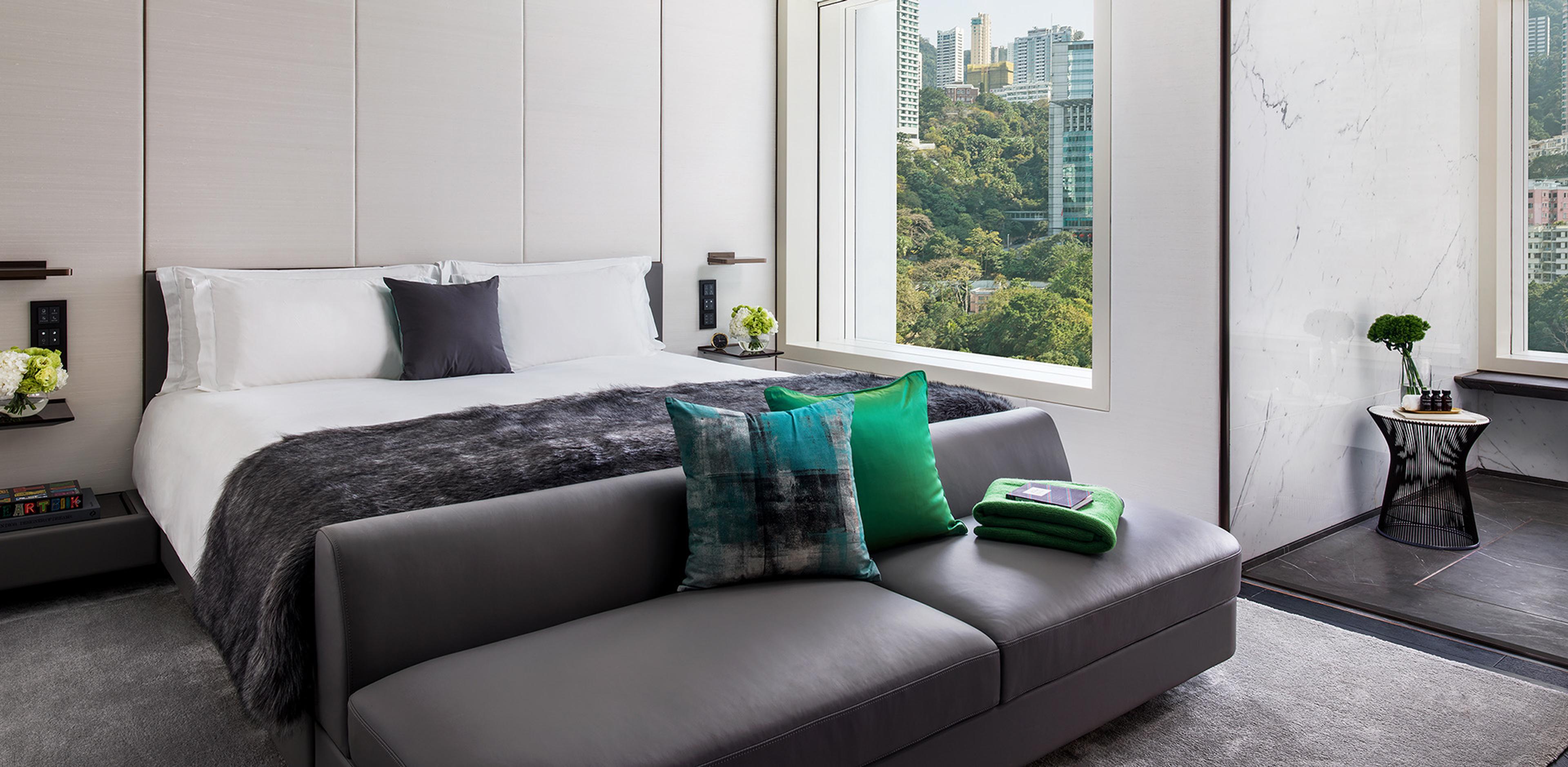 a hotel bedroom with a grey bed, a charcoal sofa with teal and green cushions, and a window framing green hillside and towers