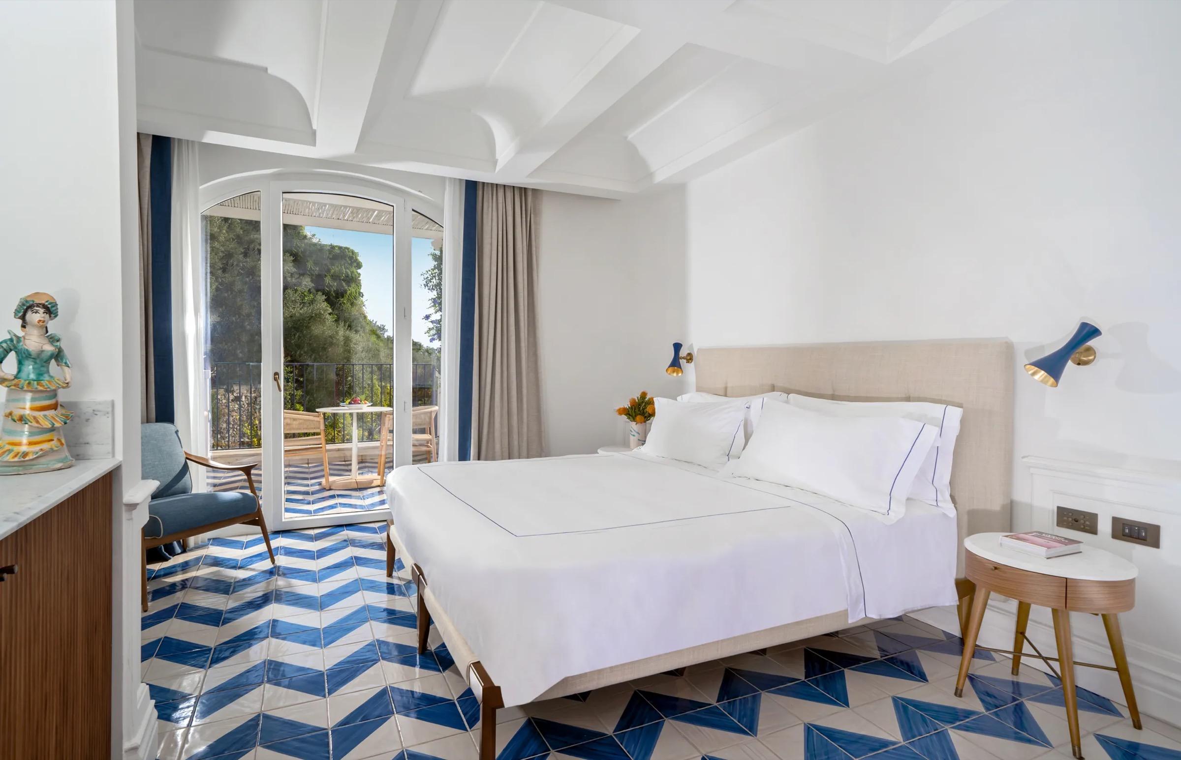 A bedroom with a chevron-patterned blue and white tiled floor, a white bed, a blue armchair, and glass doors open to a balcony over greenery.