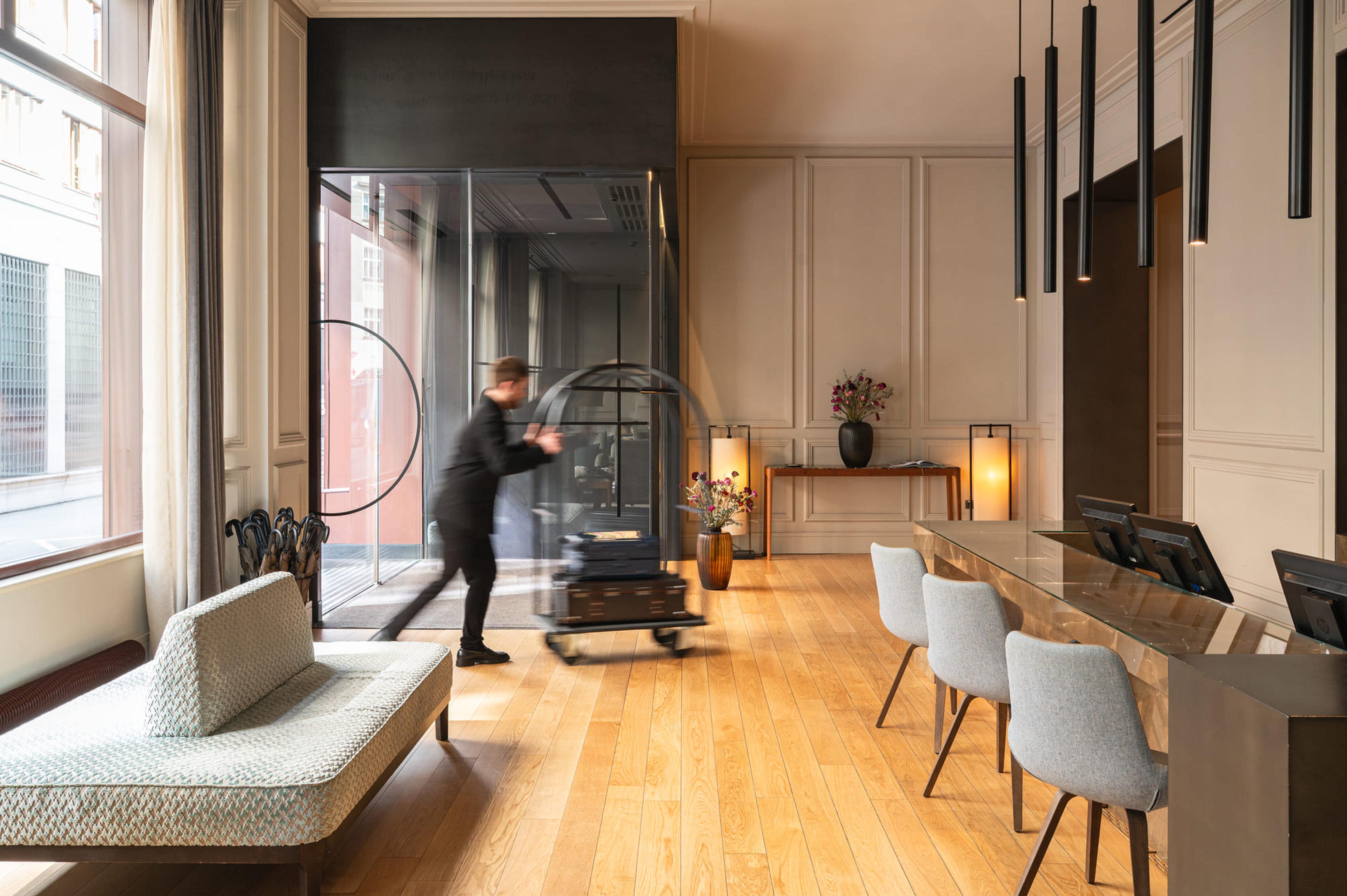 A tall reception hall with panelled walls and pendant lights, a bellhop pushing a luggage trolley across the oak floor, and a desk with screens along the right.