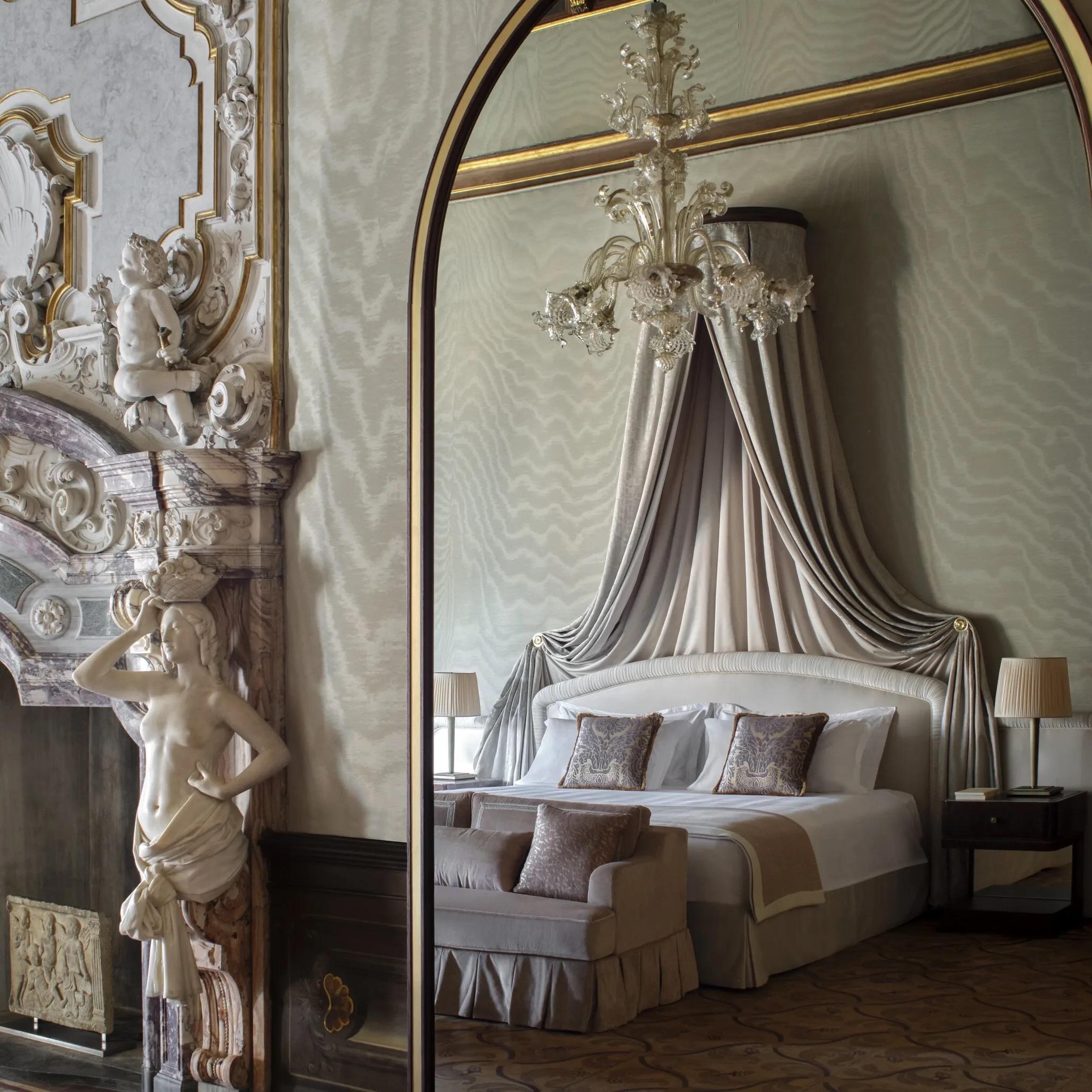 A bedroom corner with a carved marble fireplace topped by a cherub relief, beside a tall mirror reflecting a canopied bed draped in pale silk under a chandelier