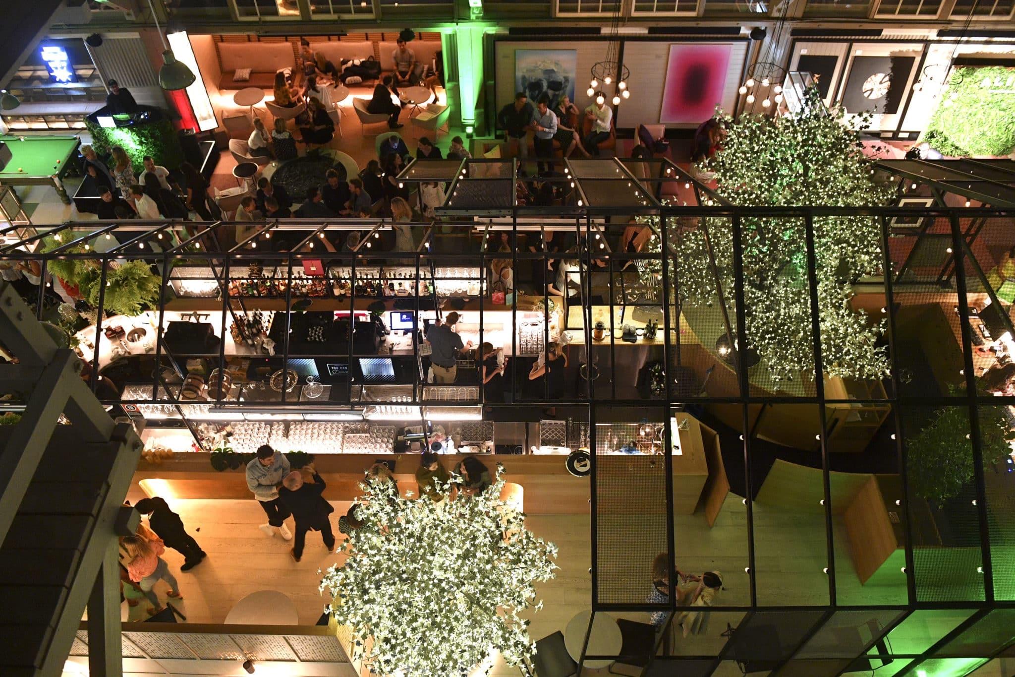 An aerial view at night of a glass-roofed courtyard bar with illuminated trees, a lit bar counter and groups of people at tables and on the floor below.