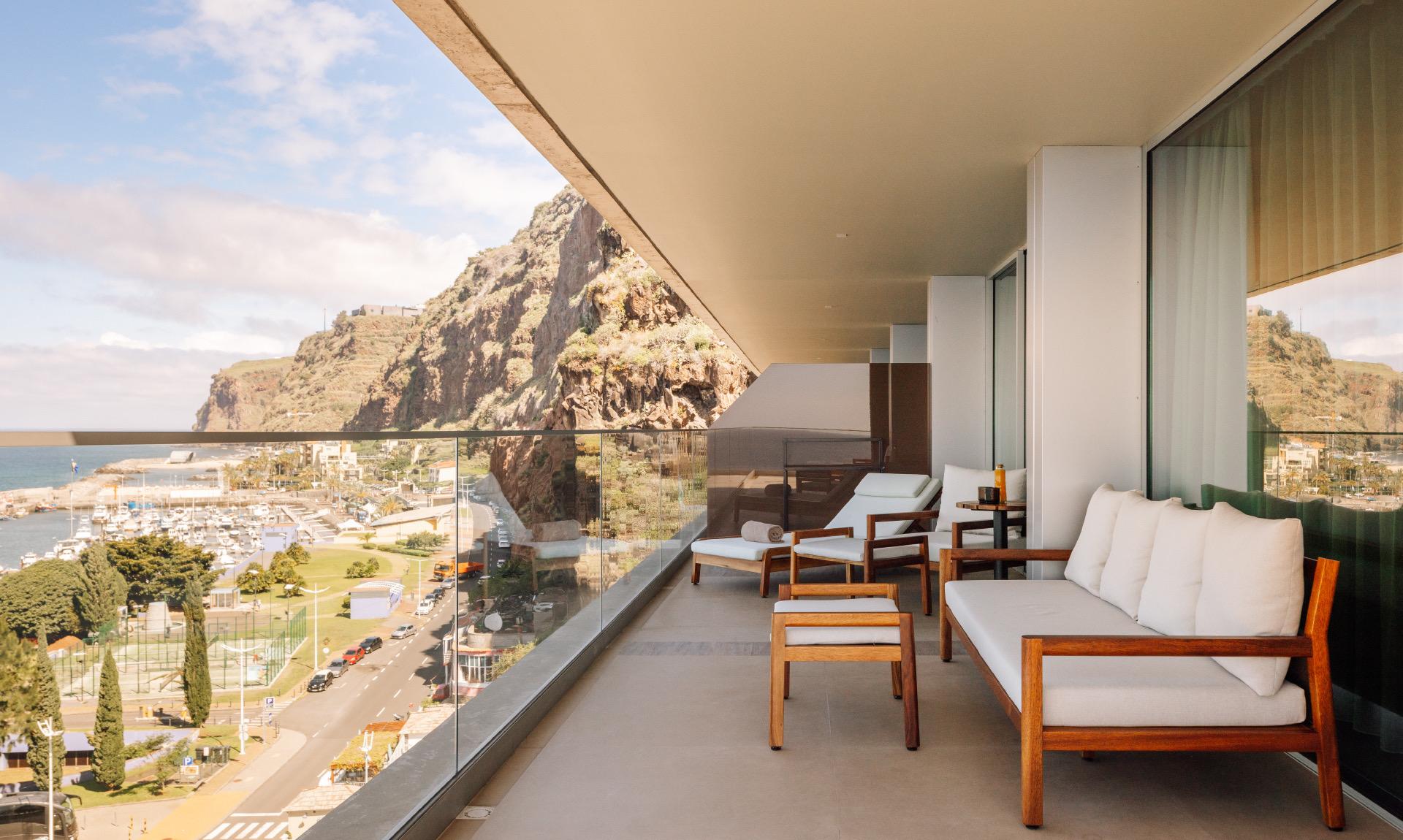 A balcony with wooden furniture looking down over a coastal town, a marina and a headland.