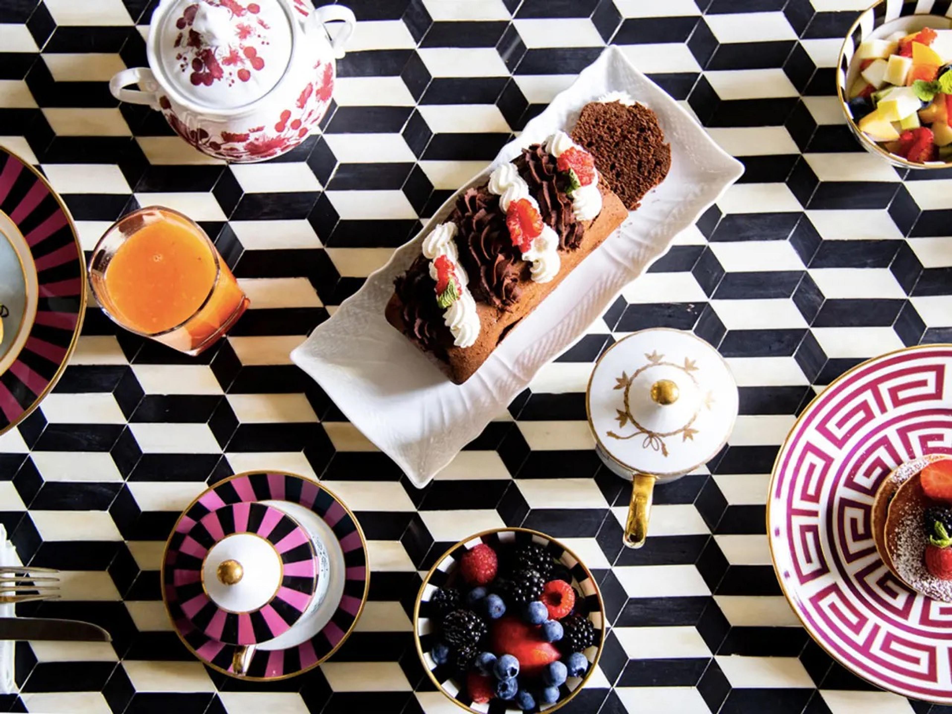 A black and white geometric-tiled table set with a chocolate loaf cake topped with cream and strawberries, a bowl of mixed berries, fruit salad, orange juice, and patterned pink and white china.