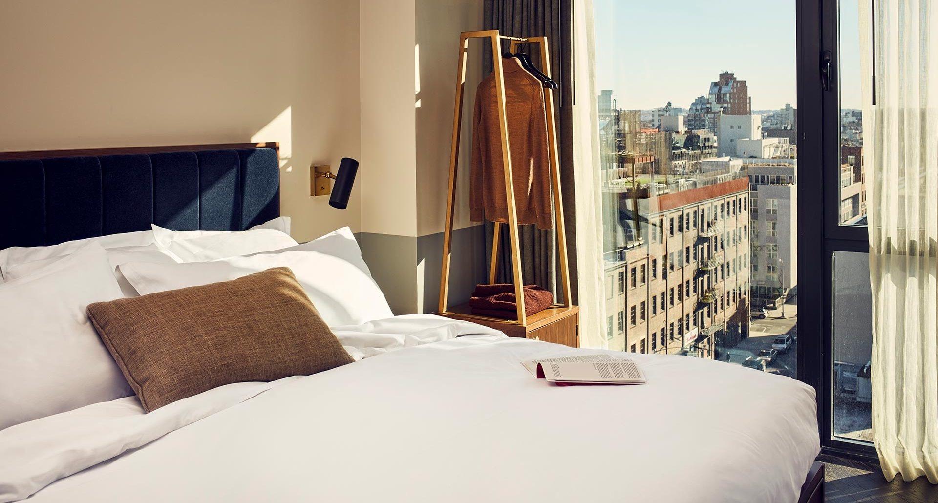 a bed with a brown pillow and a clothing rail holding a rust-colored sweater, beside a window showing a dense row of city buildings