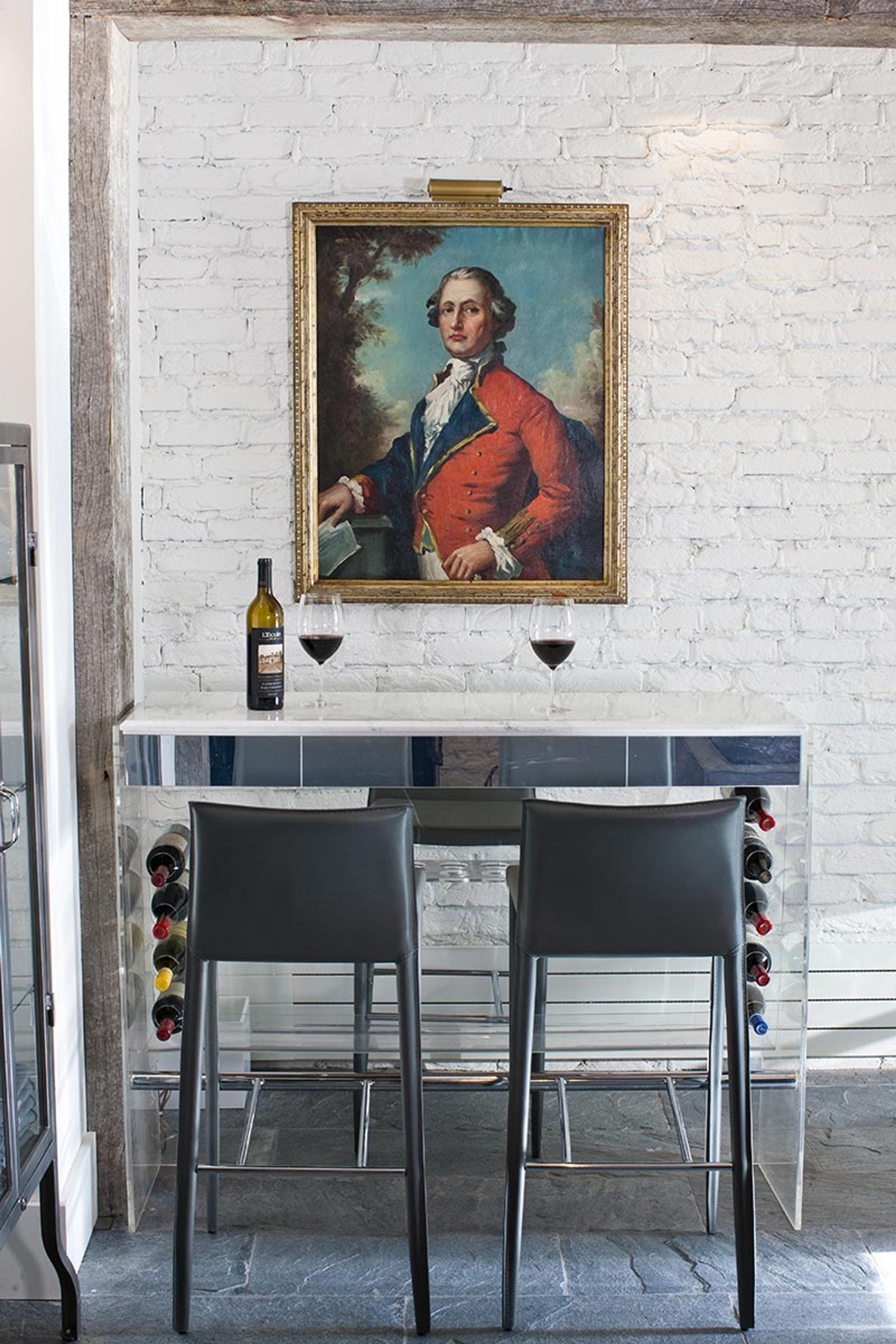 A small bar nook with a wine bottle and two poured glasses on a marble counter, under a framed portrait on a whitewashed brick wall.