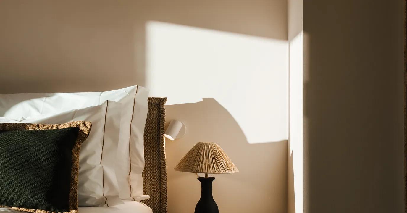 A wicker bed headboard with white linens edged in dark piping and a black cushion, a raffia-shaded lamp on the nightstand, and a sharp band of sunlight crossing the wall above.