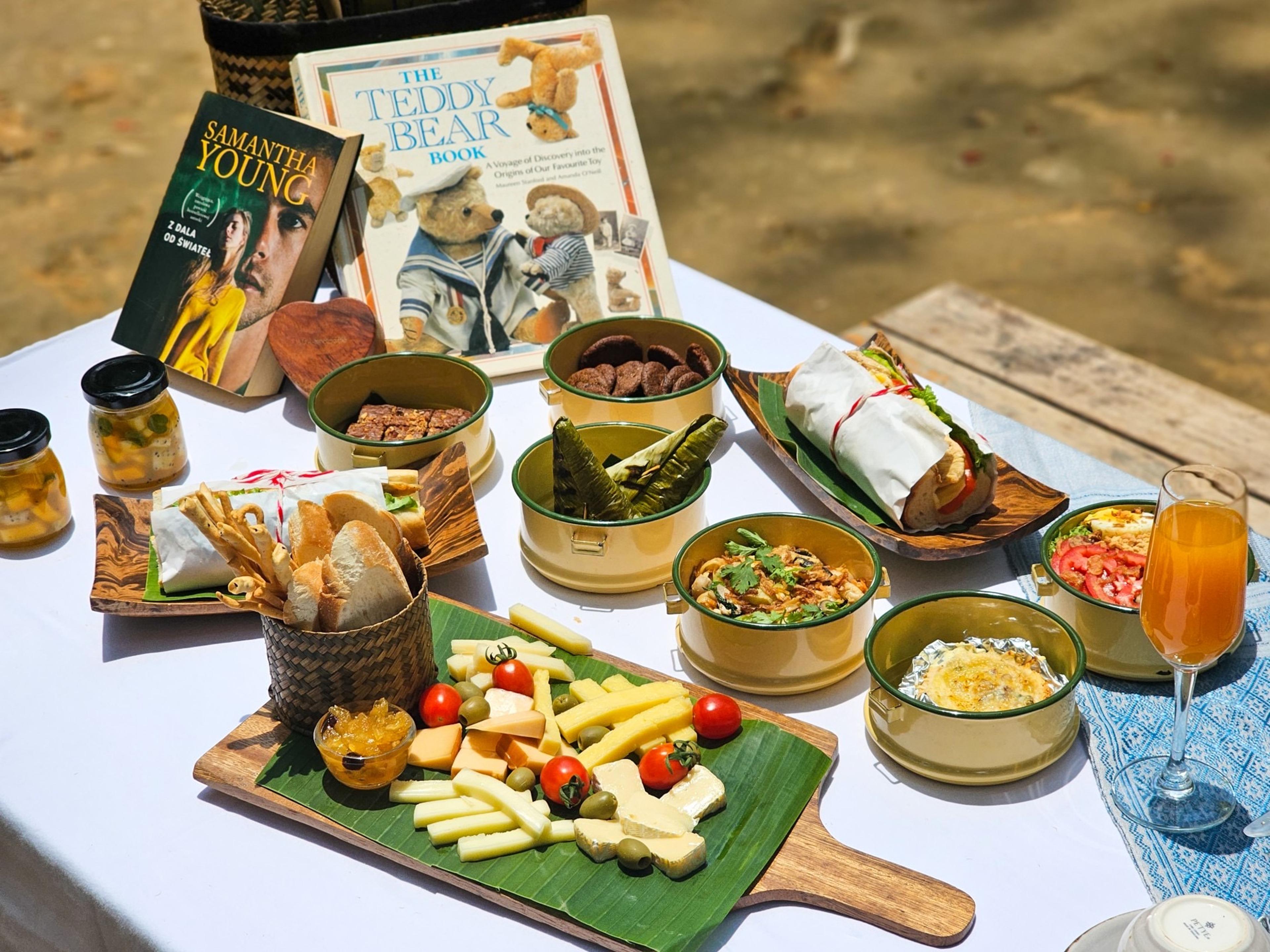 An overhead view of a picnic spread on a white tablecloth, with bread, cheese, charcuterie, pickles and a wrapped sandwich, and two books propped against a basket.