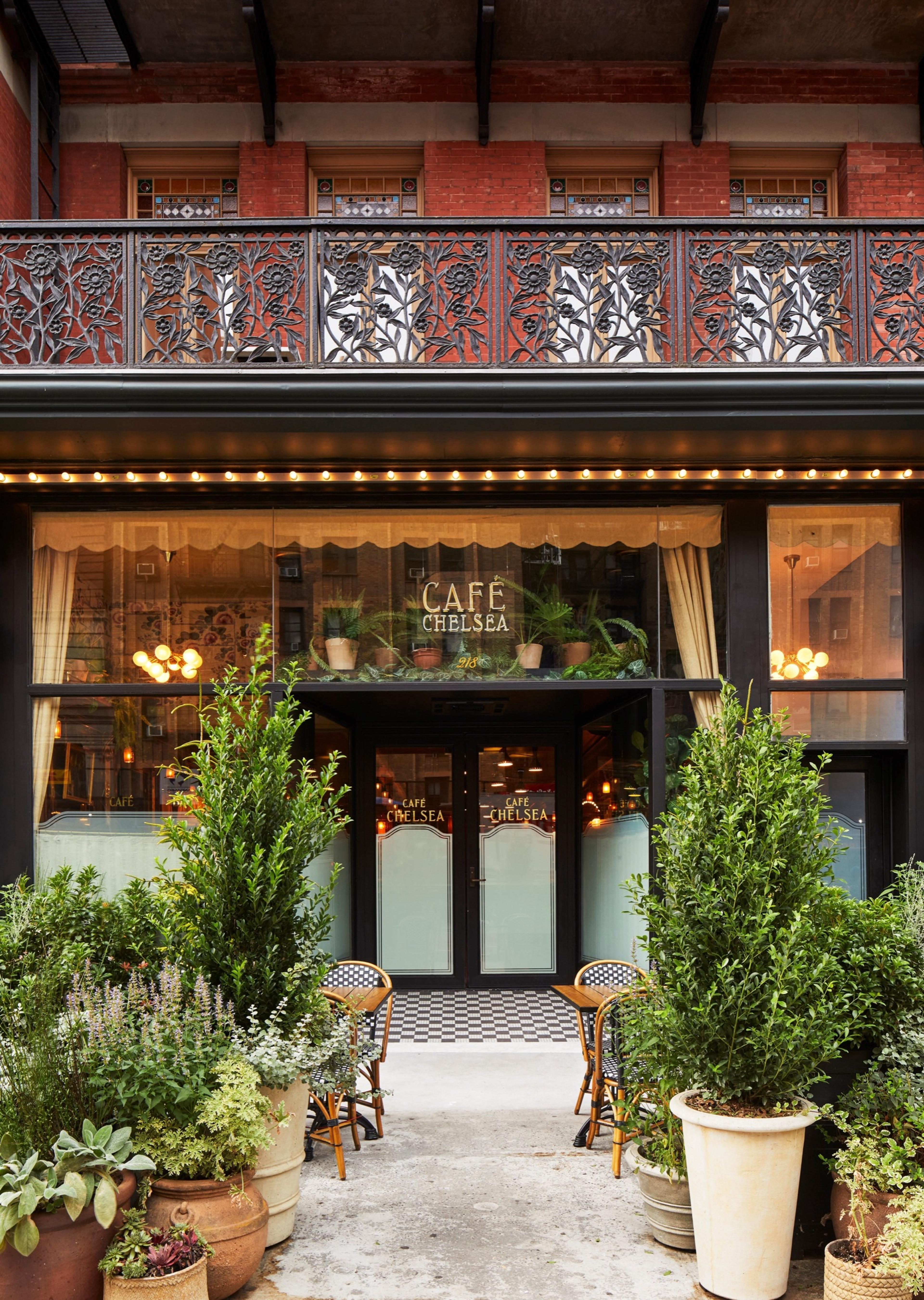 The street level entrance to Cafe Chelsea, with glass doors, a lit awning, potted topiary, and an ironwork balcony above.