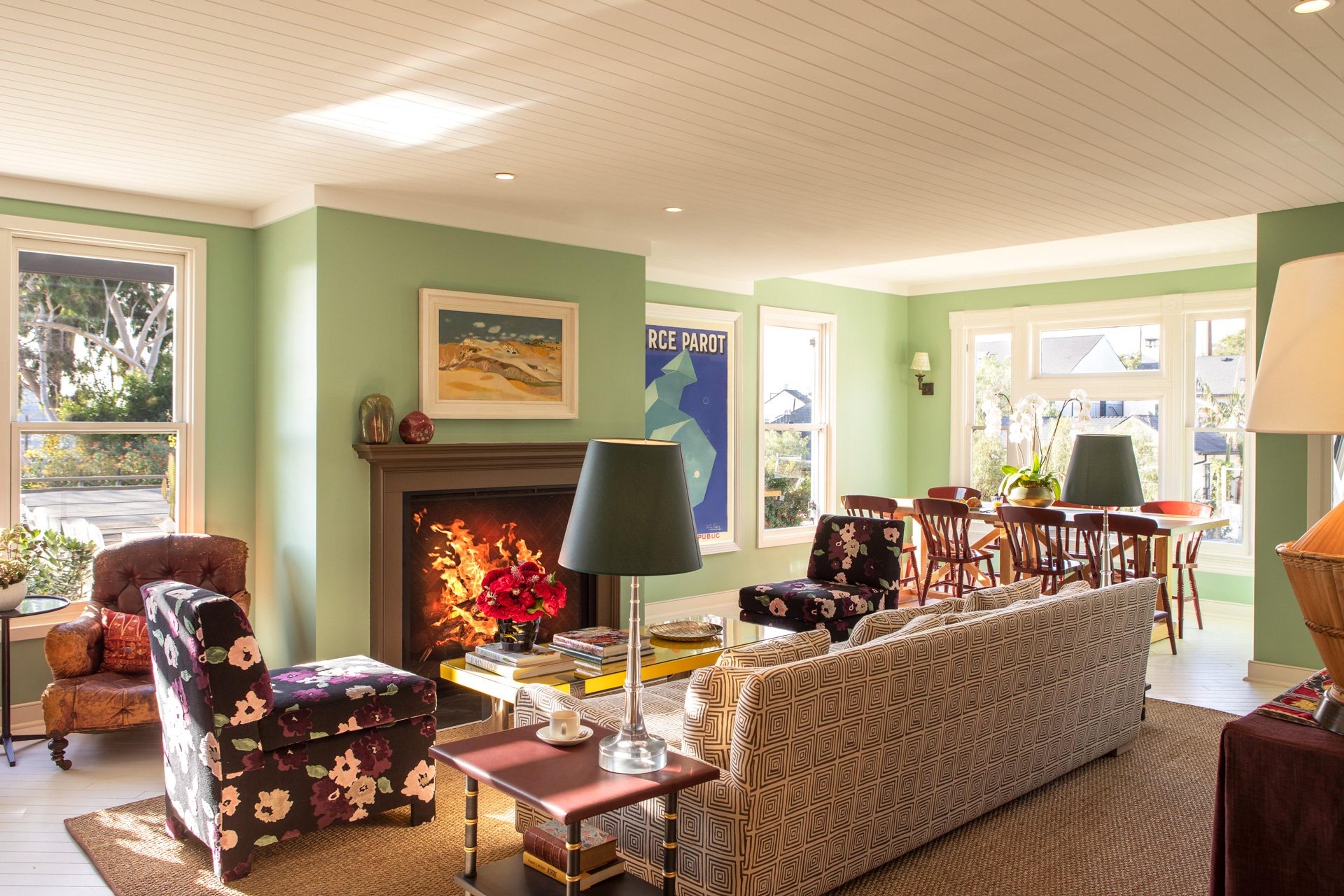 A bright living room with pale green walls, a lit fireplace, patterned armchairs and a sofa, and a dining table and chairs visible beyond.