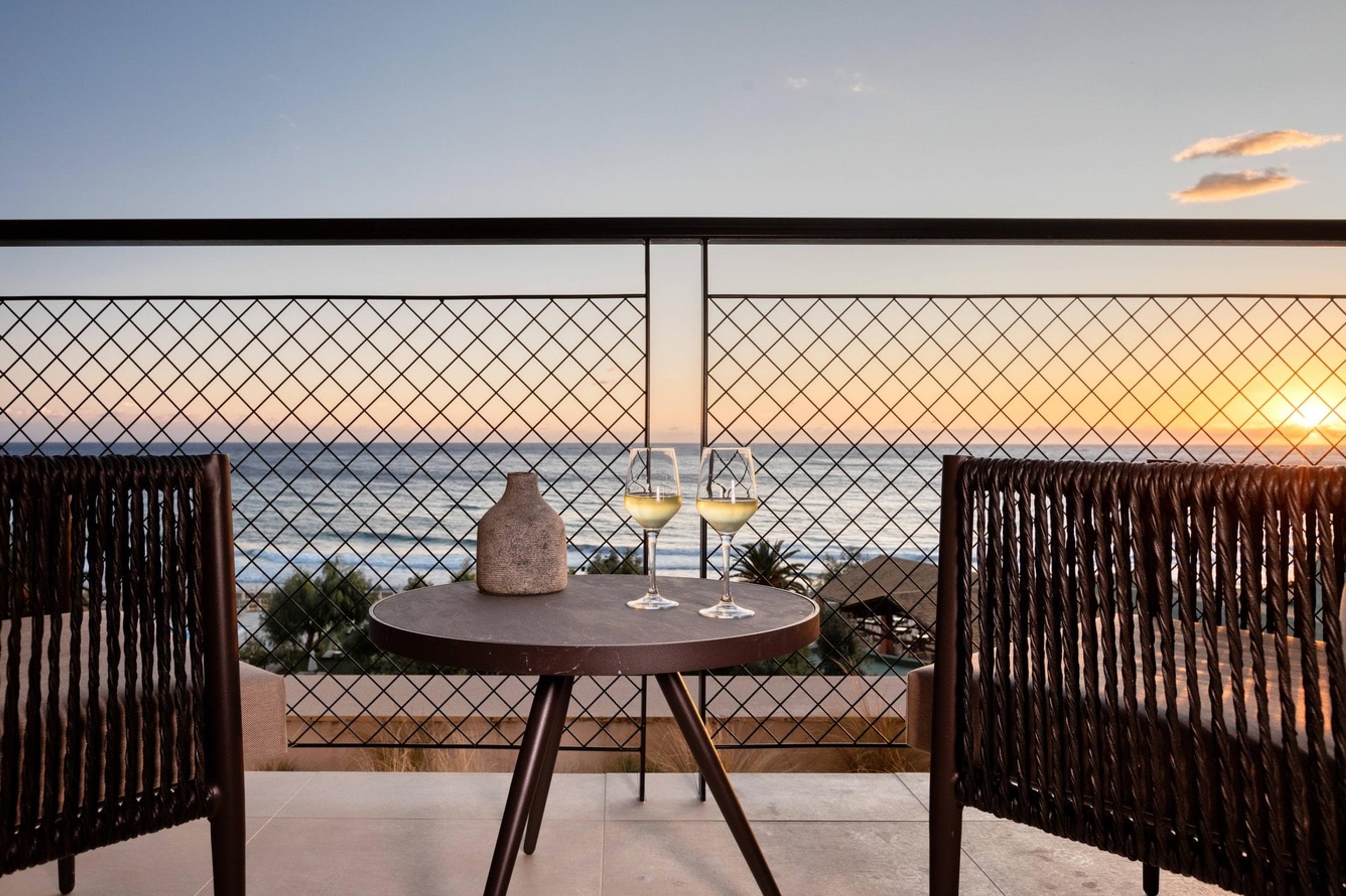 Two glasses of white wine on a balcony table between woven chairs, with the sun setting over the sea through a diamond-patterned railing.