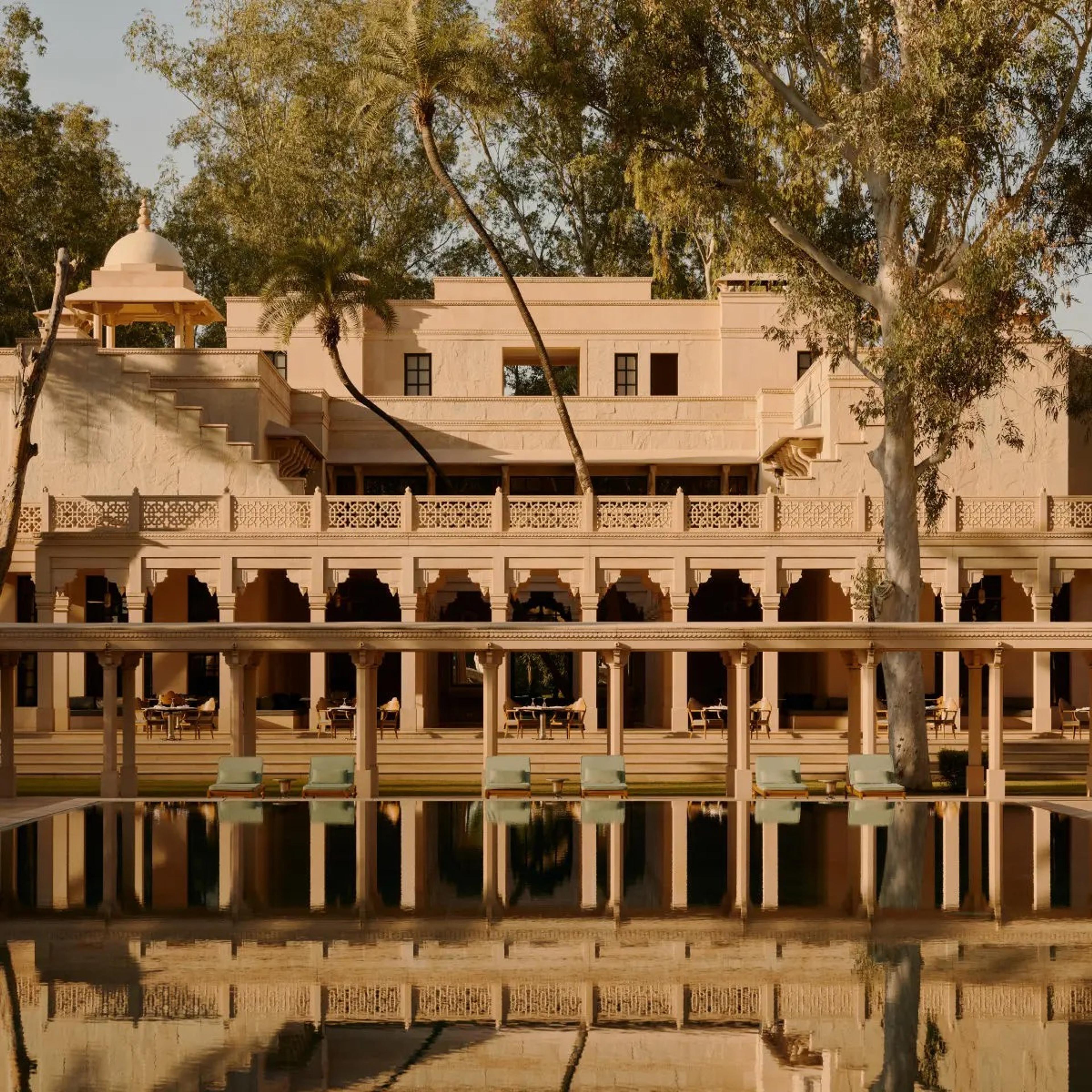 A reflecting pool stretching along a cream stone colonnade and sun terrace, with green loungers under palm trees and a domed pavilion behind.