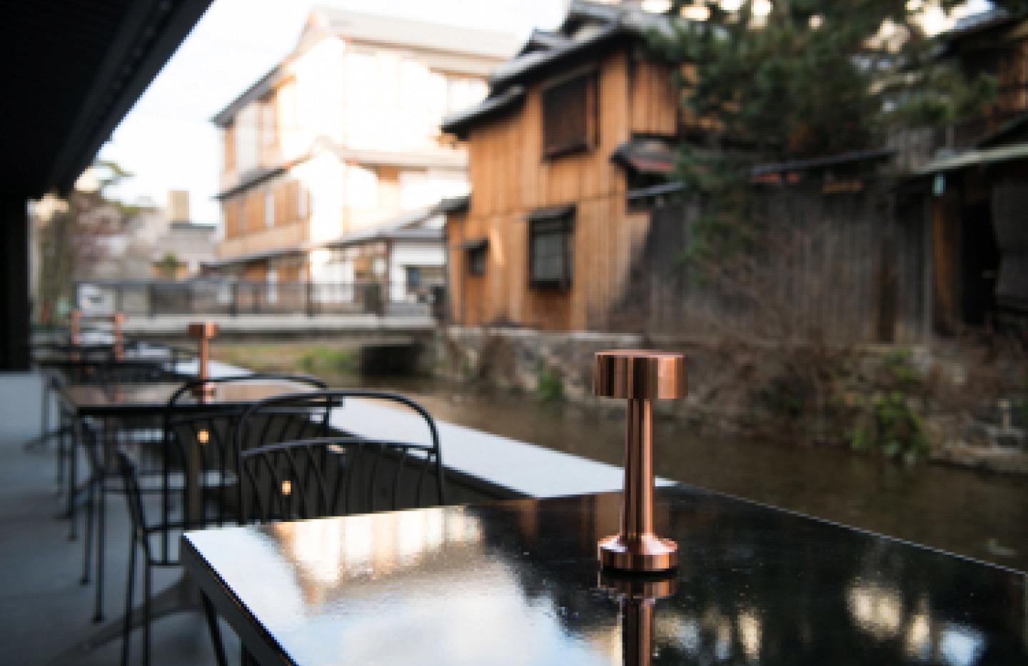 outdoor dining tables along a canal in daylight, with traditional wooden houses on the opposite bank