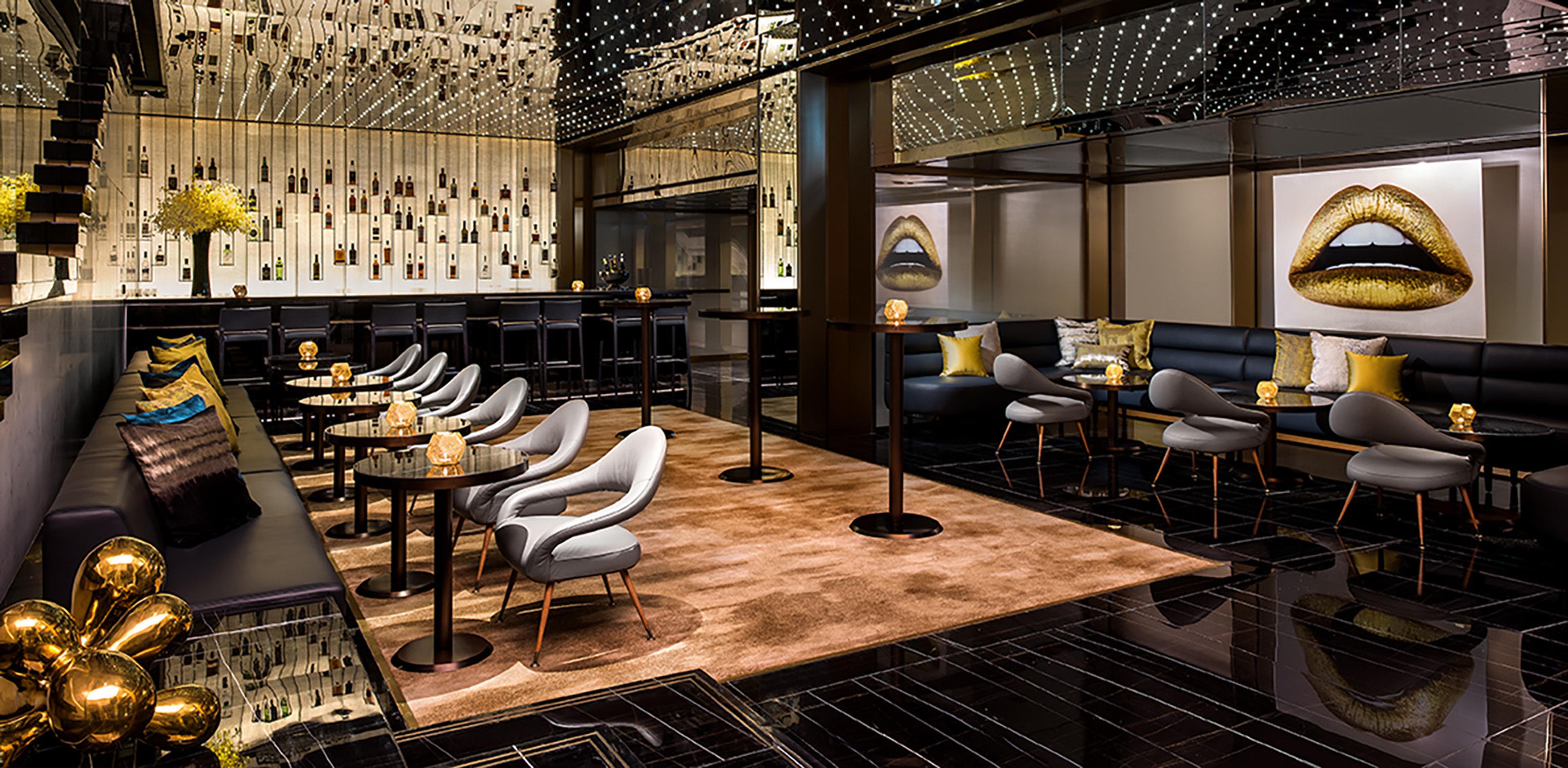 a hotel bar and lounge with grey armchairs, a backlit bottle wall and a gold toned artwork of lips on the far wall