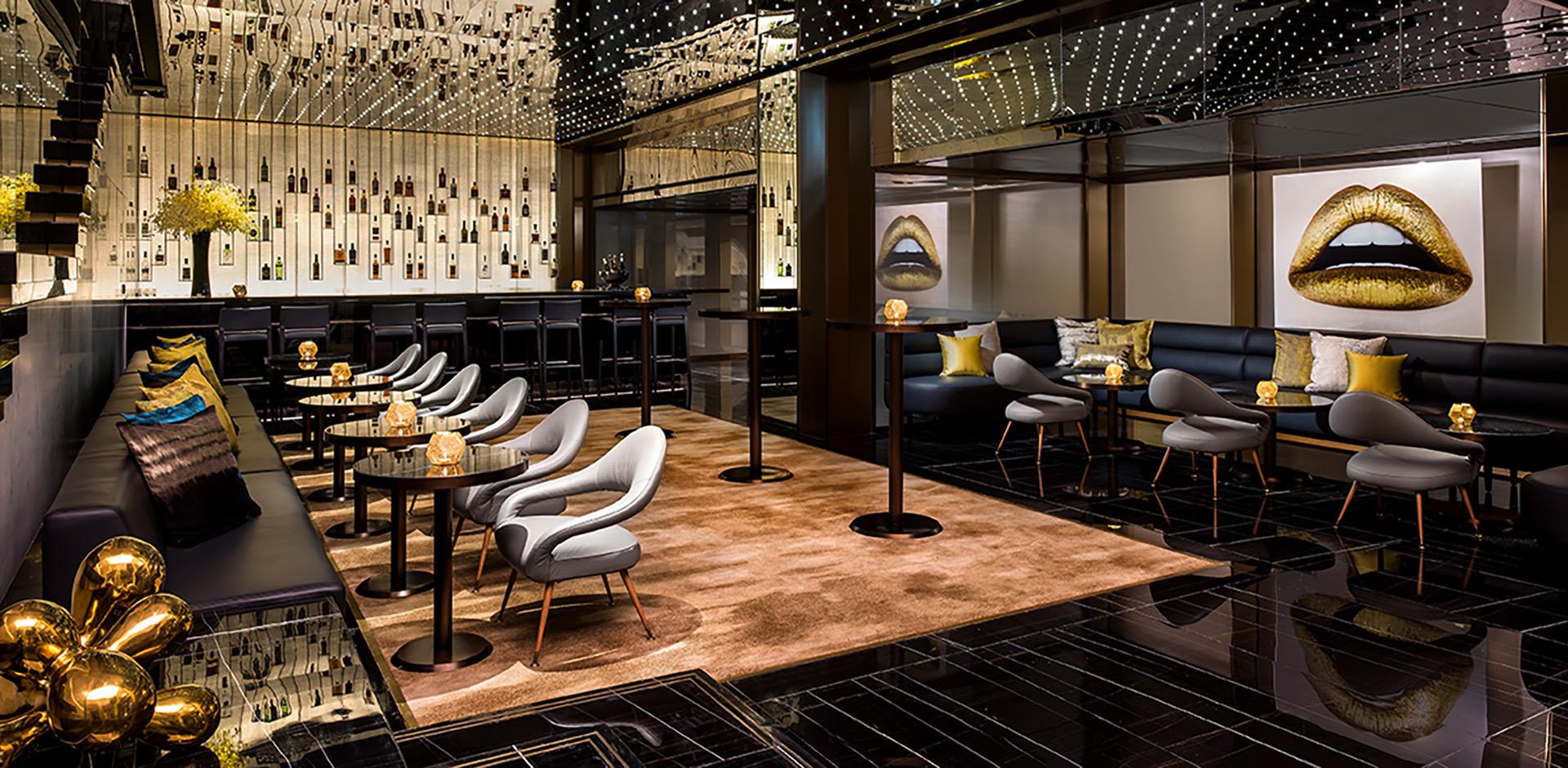 a hotel bar and lounge with grey armchairs, a backlit bottle wall and a gold toned artwork of lips on the far wall