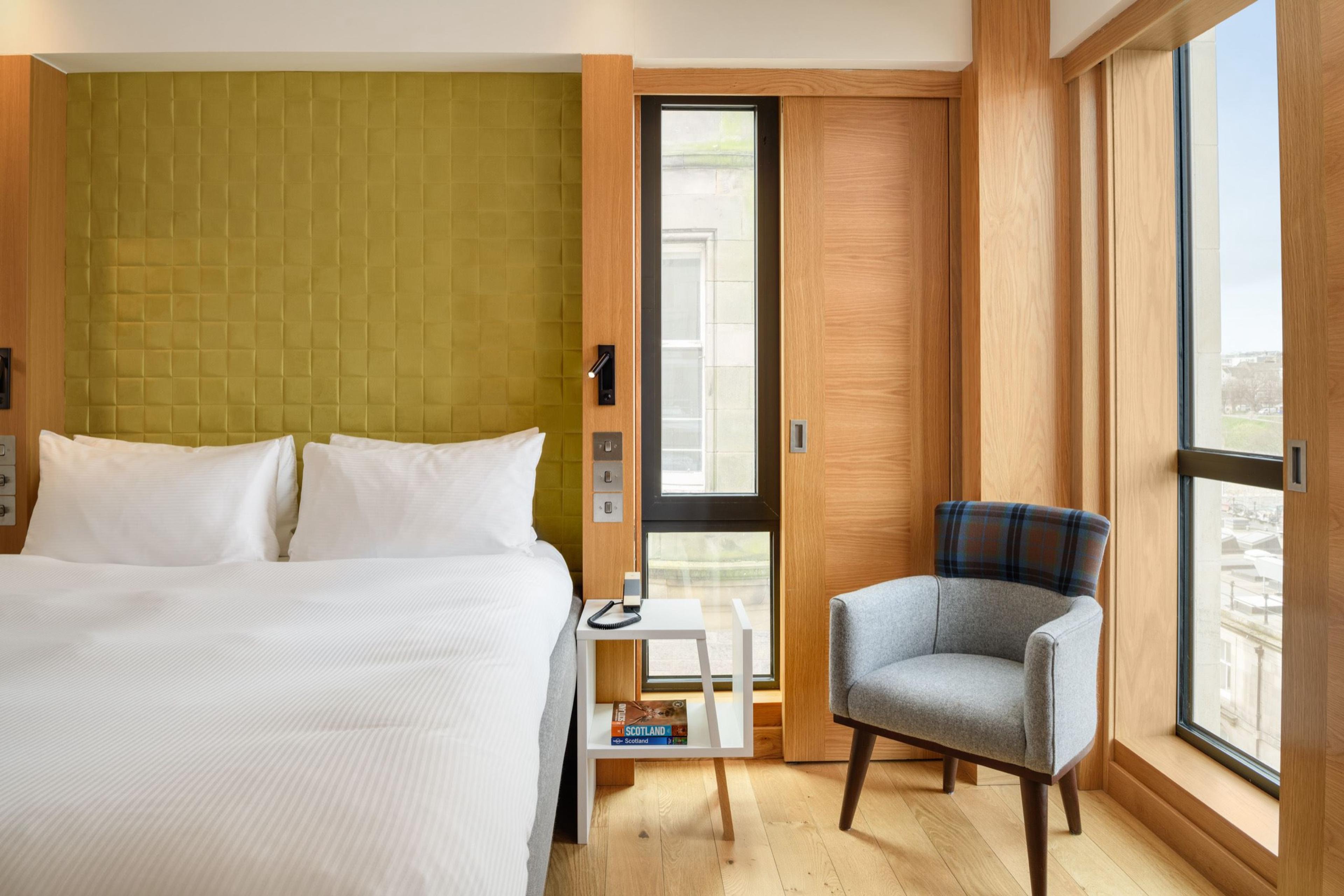 A bed with a green textured headboard in a wood-panelled room, with a tartan armchair and two tall windows.