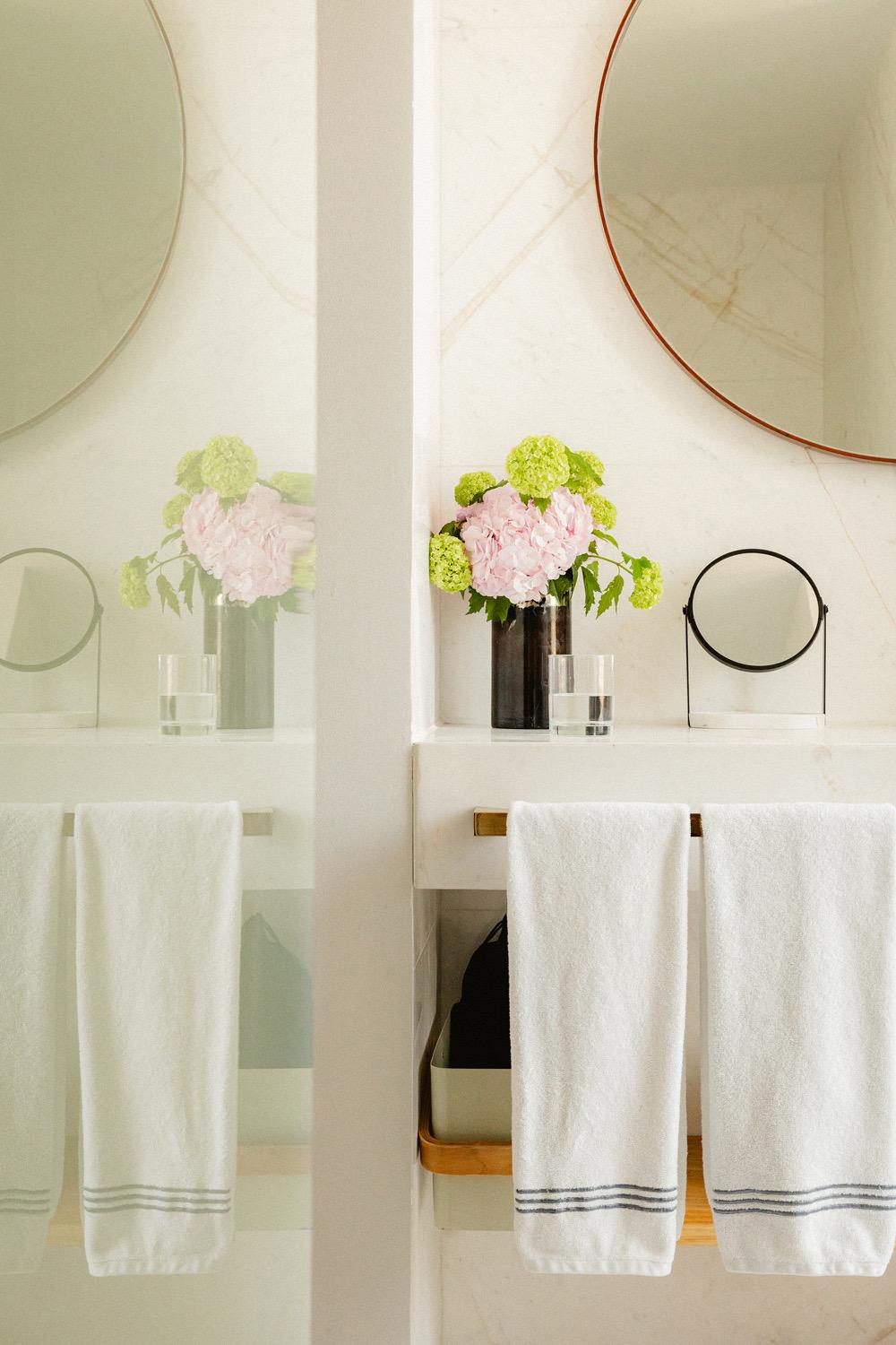 Two bathroom counters, each with a round mirror, a vase of pink and green flowers and folded white towels.