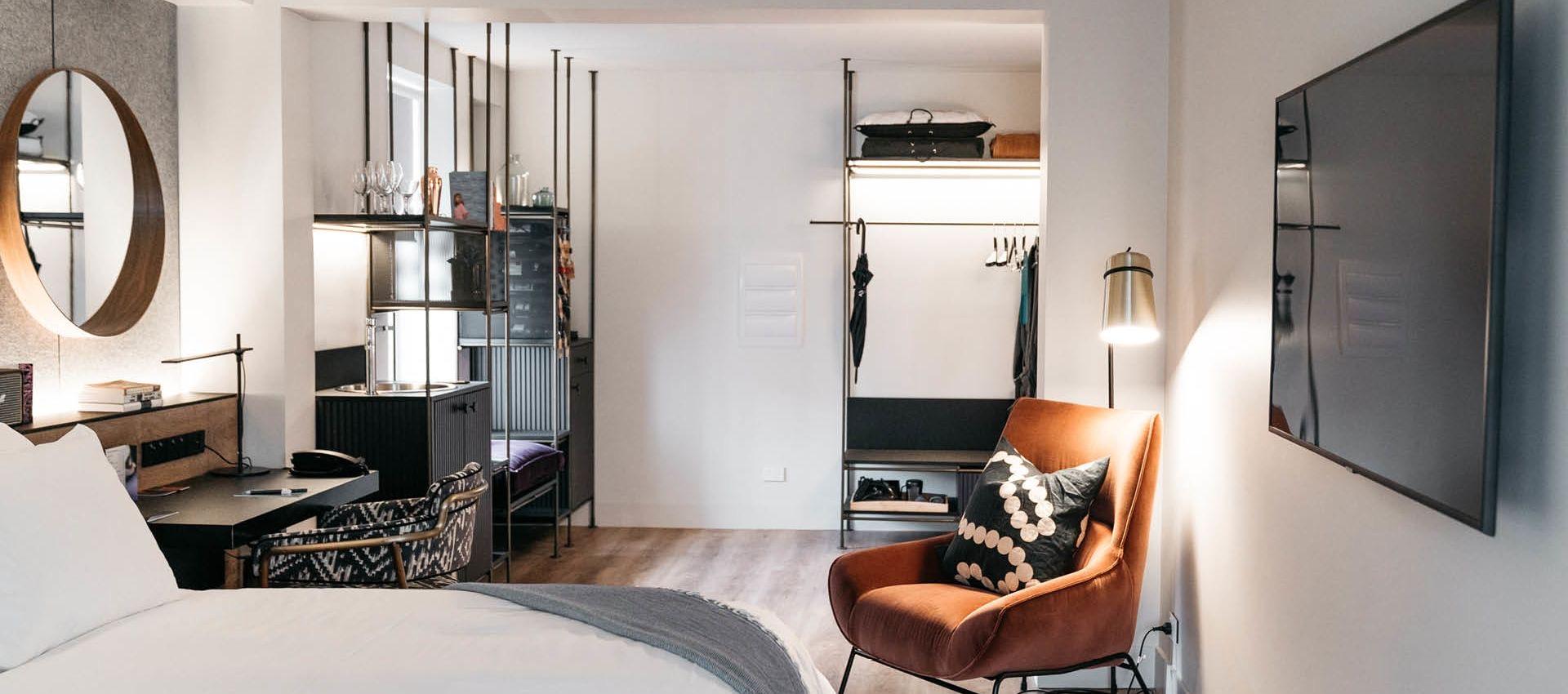 a hotel suite with an open wardrobe and shelving, a round mirror over the desk, and an orange armchair by the television