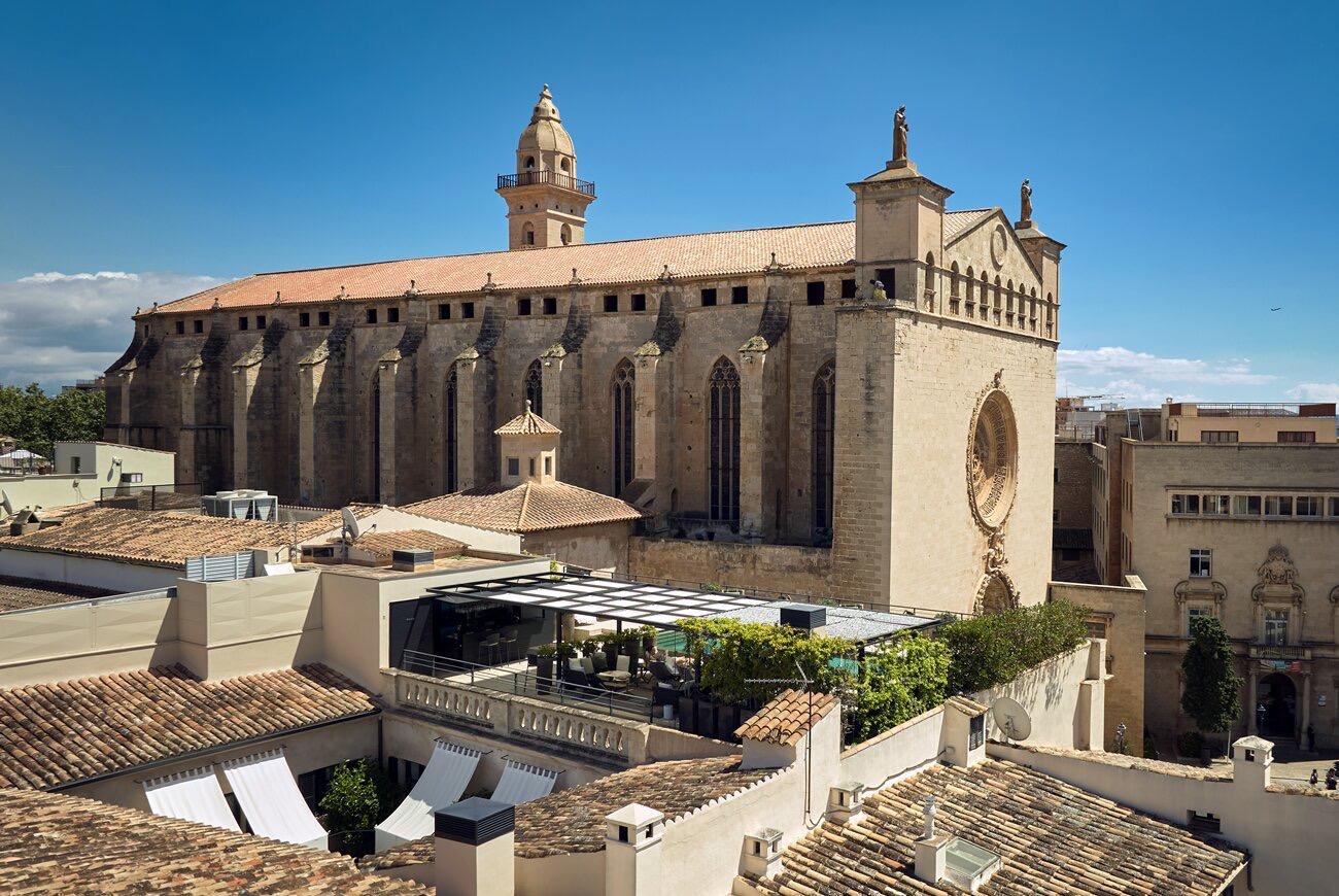 An aerial view of a Gothic church's stone flank, its rose window and bell tower rising above surrounding rooftops, with a terrace of white umbrellas and a strip of swimming pool in the foreground.