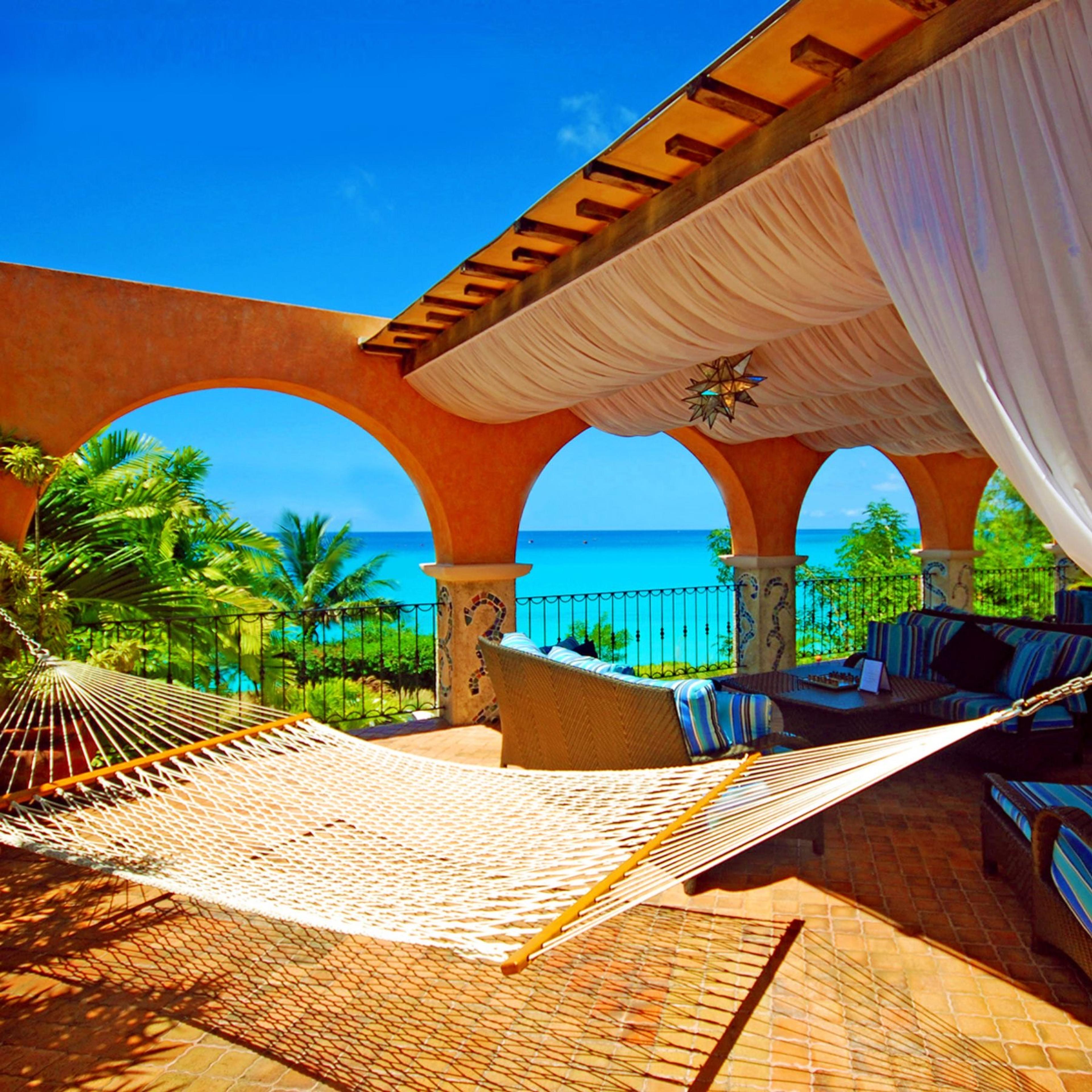 A terrace with a hammock strung between stone pillars, wicker furniture, and a view of the sea through a row of arches.