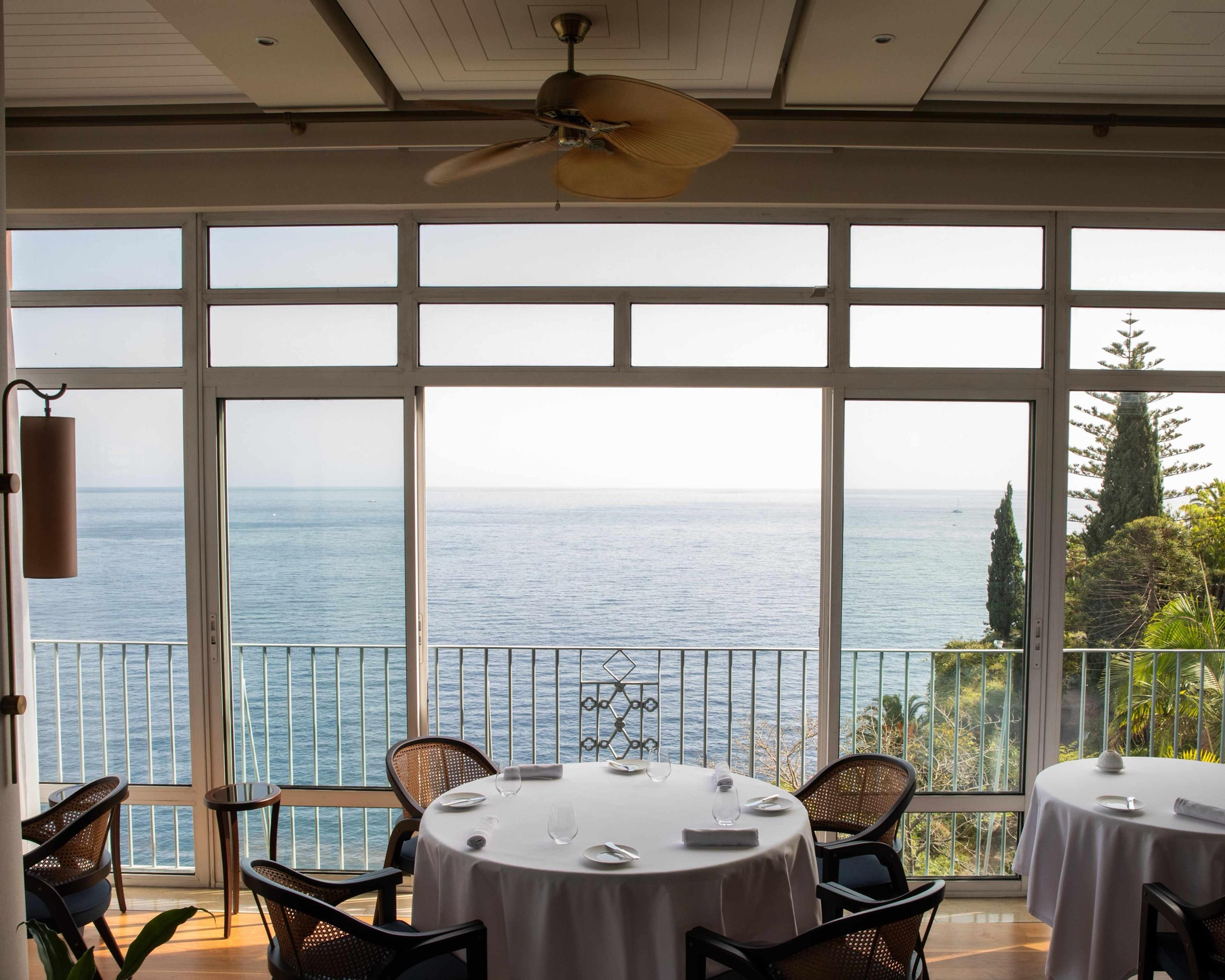A ceiling fan hangs over a restaurant with floor-to-ceiling windows, round tables laid with white cloths and glassware, the sea filling the view beyond.