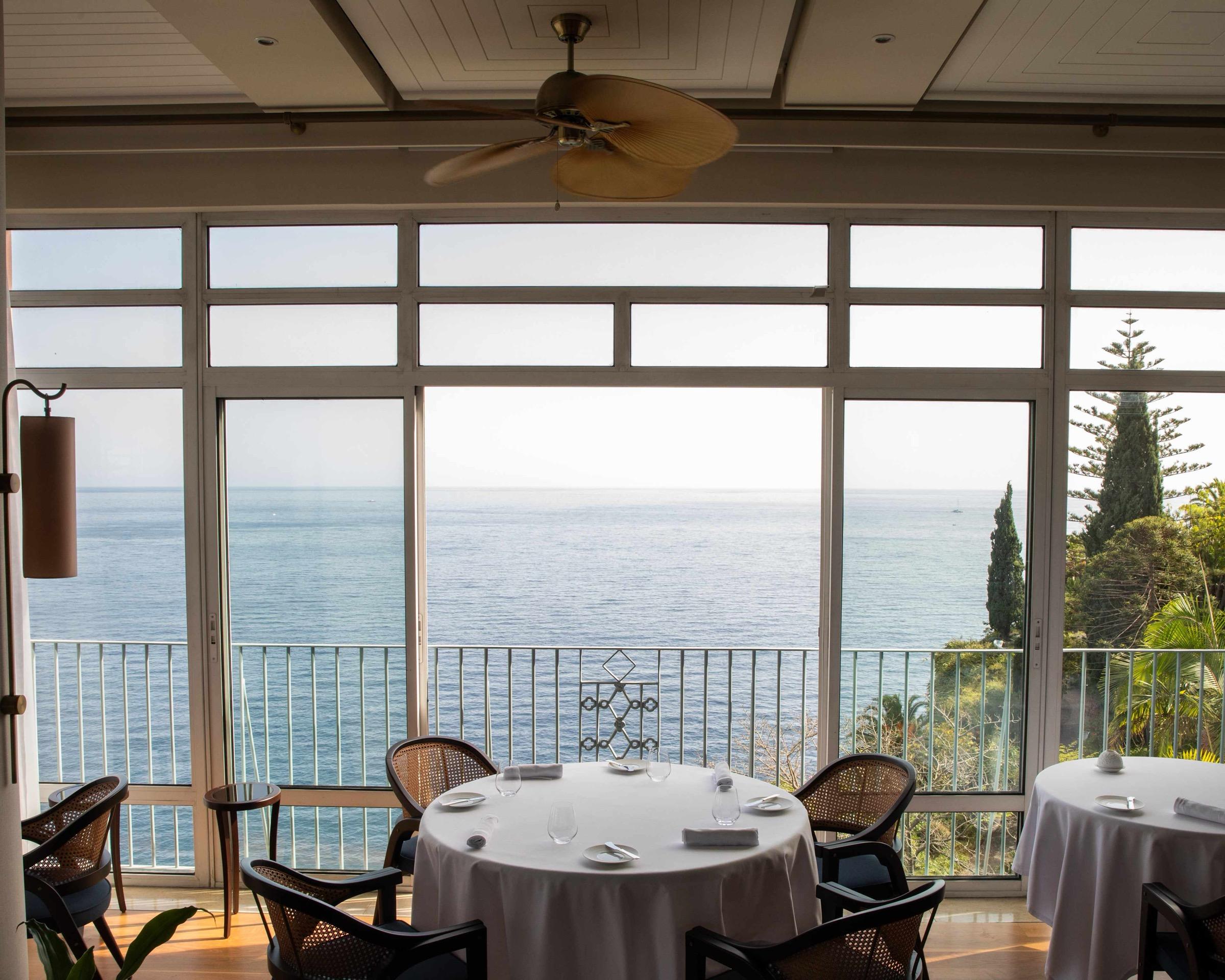 A ceiling fan hangs over a restaurant with floor-to-ceiling windows, round tables laid with white cloths and glassware, the sea filling the view beyond.