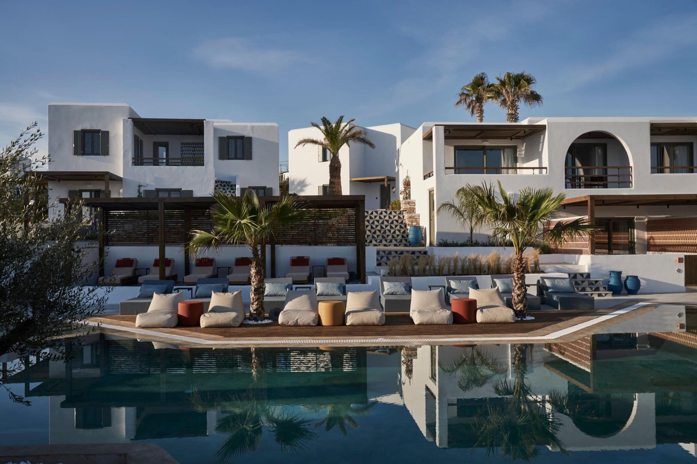 Two-storey whitewashed hotel buildings with dark-framed windows and balconies stand behind a line of palm trees and cushioned daybeds at the edge of a pool, their reflection visible in the water.