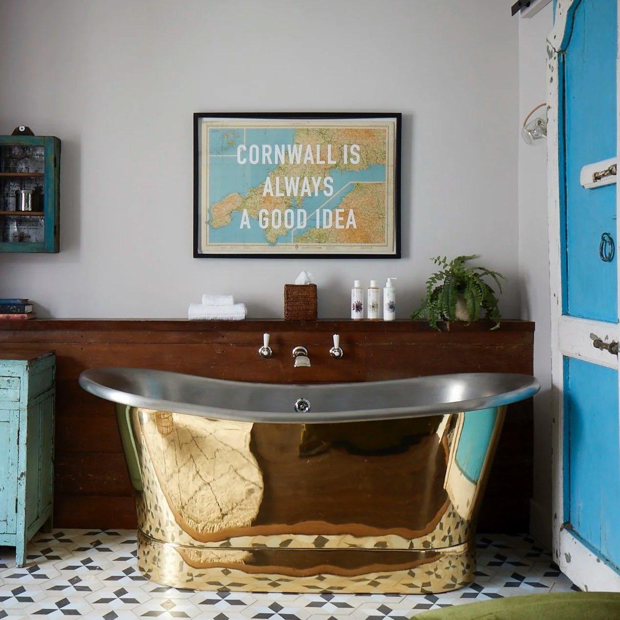 A gold and steel freestanding bathtub against a dark wood panel, below a framed print reading Cornwall is always a good idea, with a painted blue door to one side.
