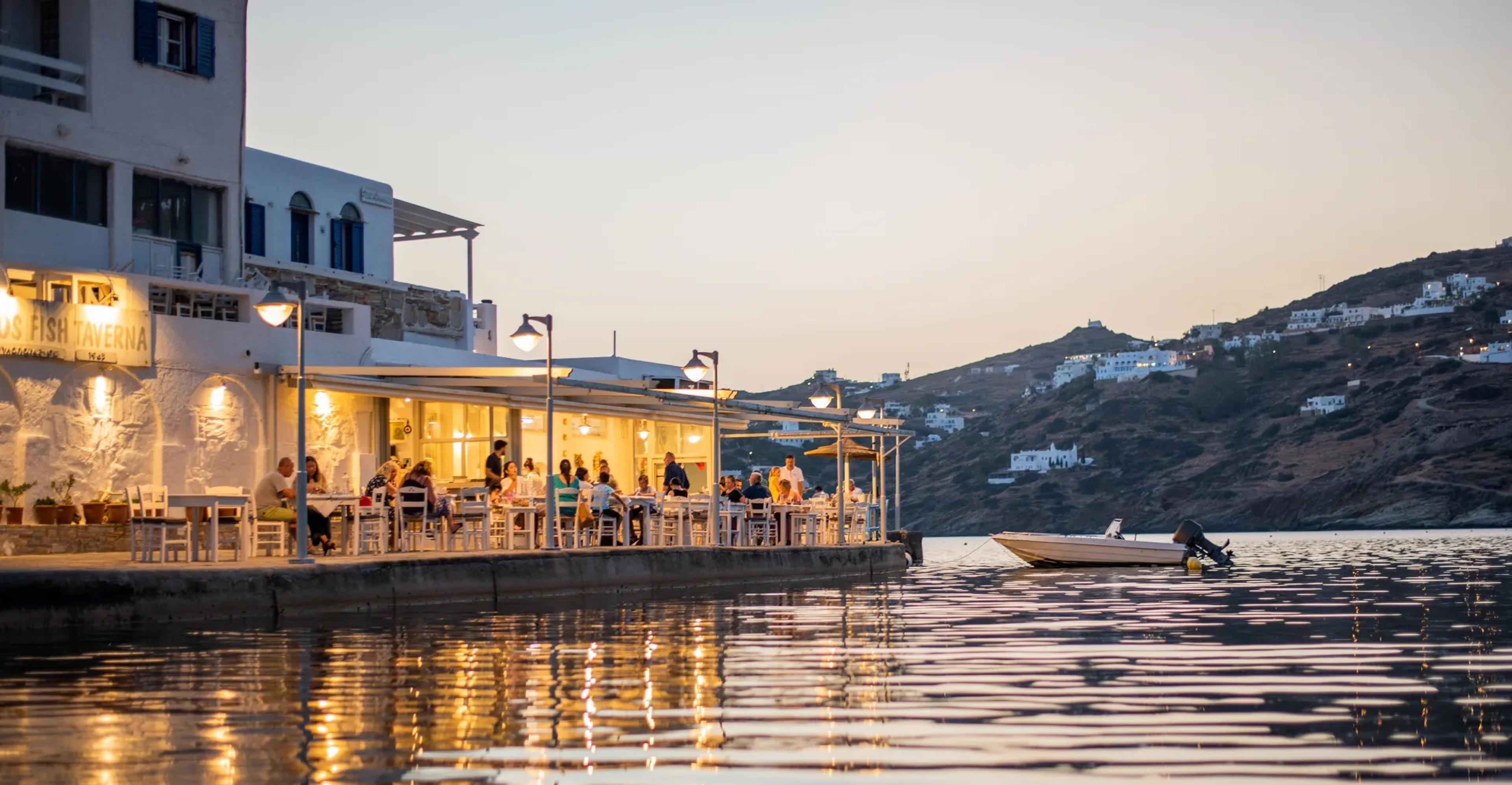 A taverna's lit tables along the water's edge at dusk, hills beyond