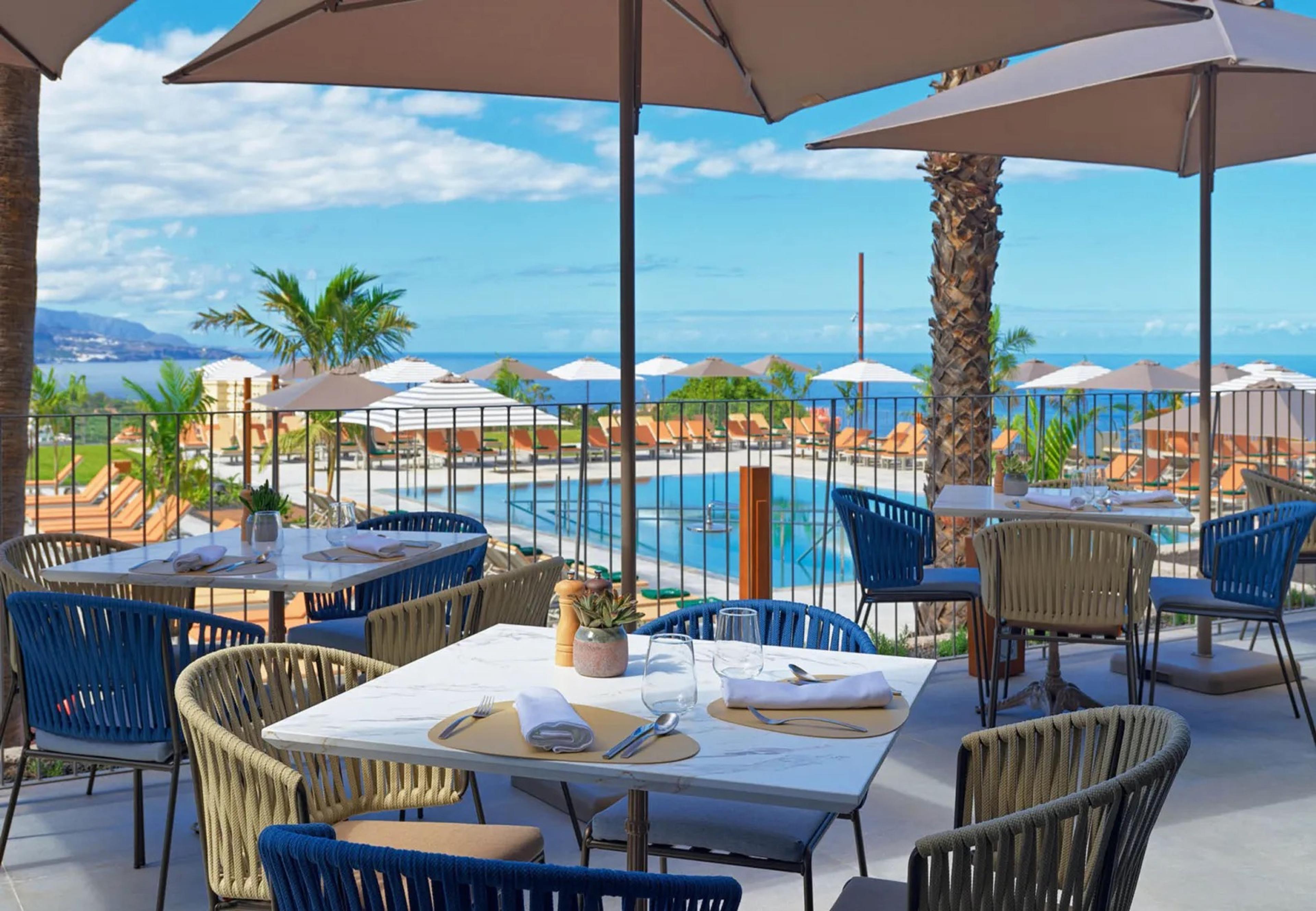 A restaurant terrace with marble tables set for lunch under umbrellas, looking over a swimming pool with loungers to the sea beyond.